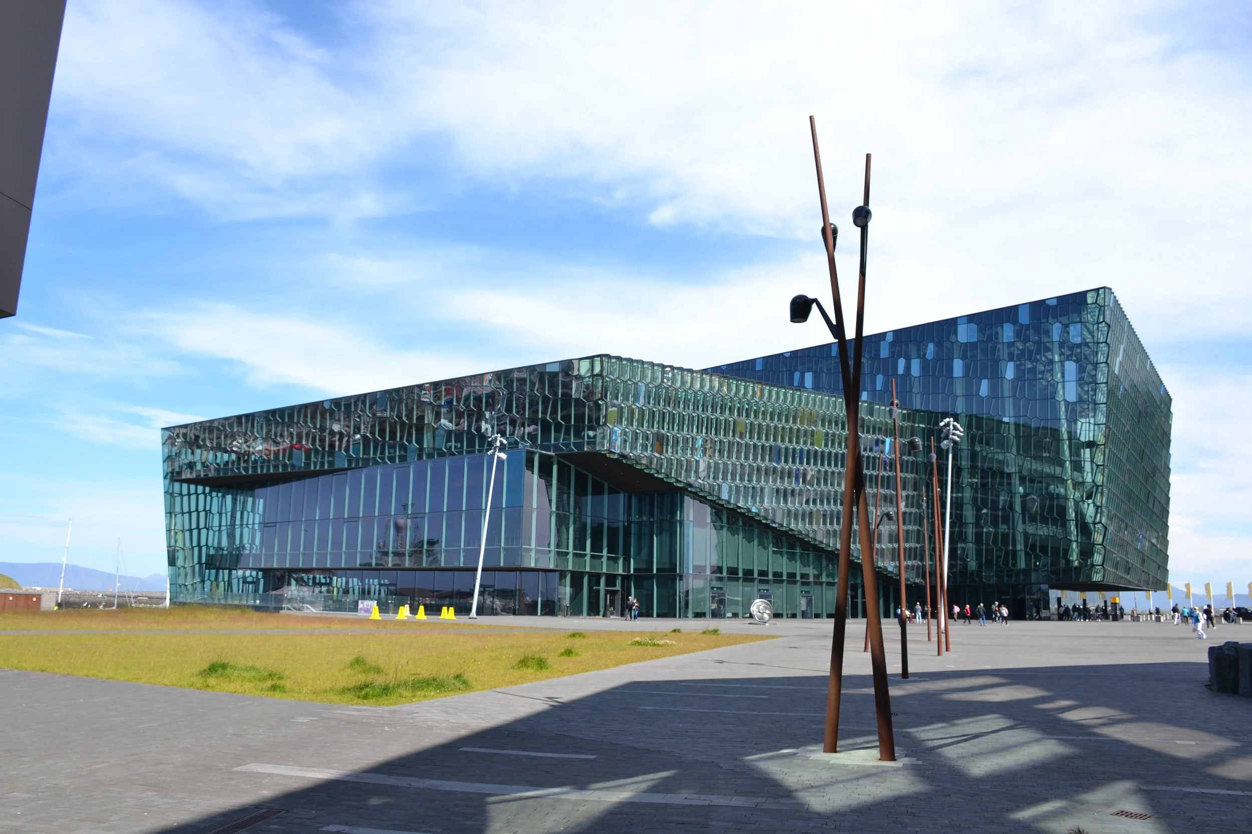Wide exterior view of Harpa Concert Hall in Reykjavík, Iceland, showing its geometric glass façade, shifting blue and green reflections, public plaza, sculptural light poles, and patterned shadows.