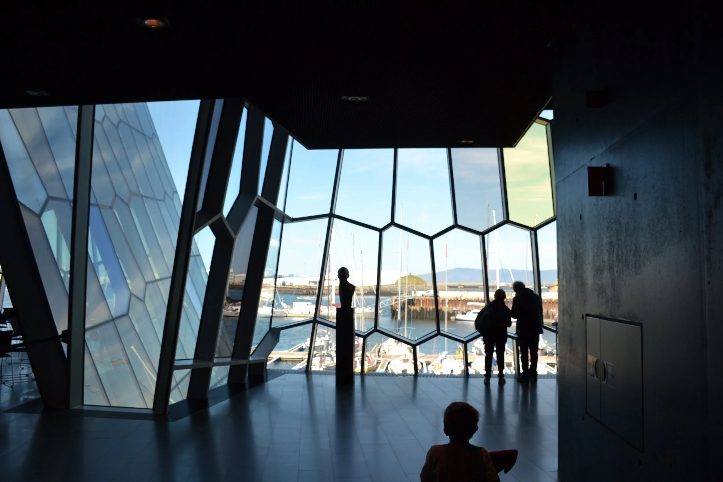 Silhouetted visitors looking across Reykjavík’s harbor through the geometric glass façade inside Harpa Concert Hall.