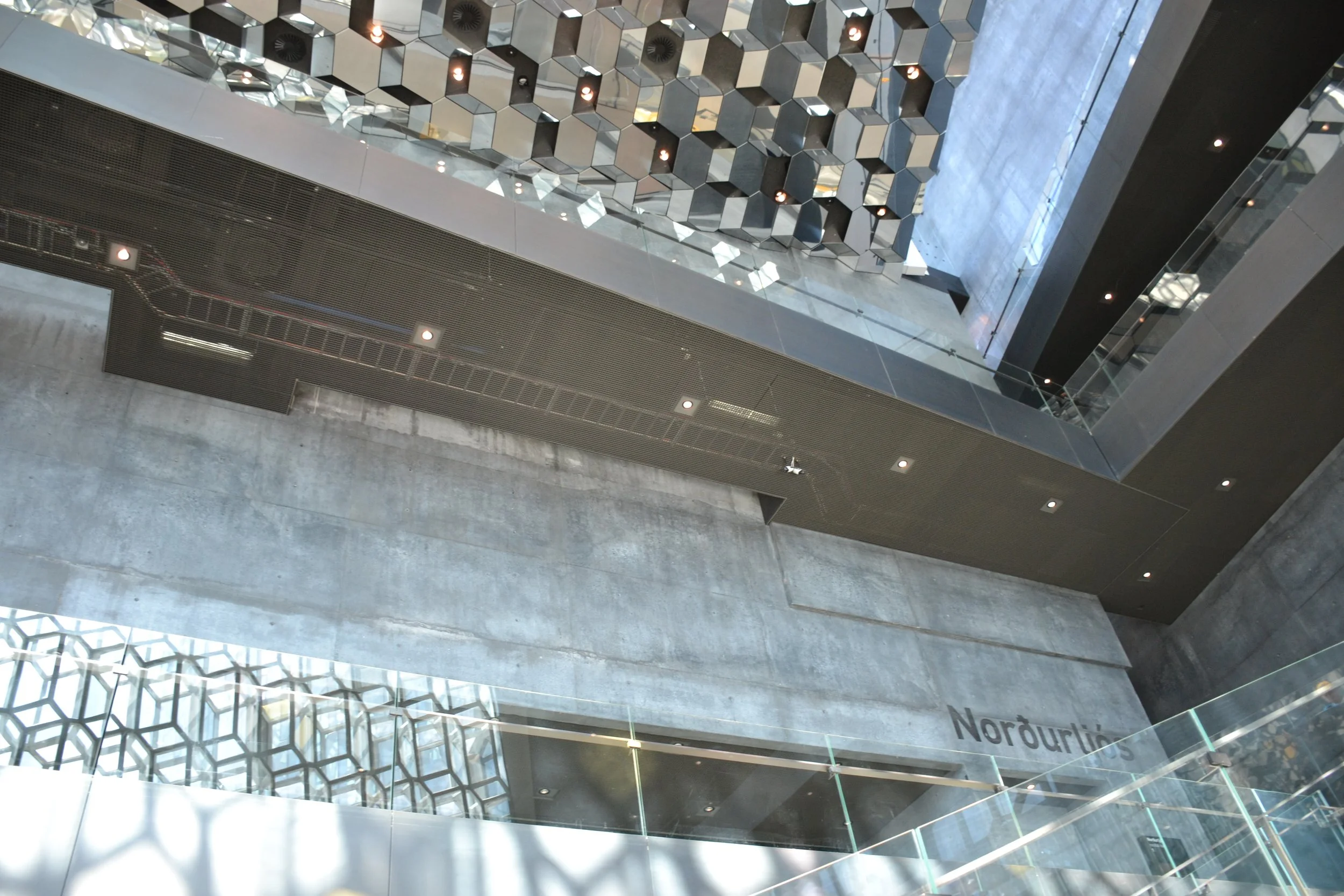 Tall interior foyer at Harpa Concert Hall with a geometric mirrored ceiling, glass façade, dark walls, and daylight.