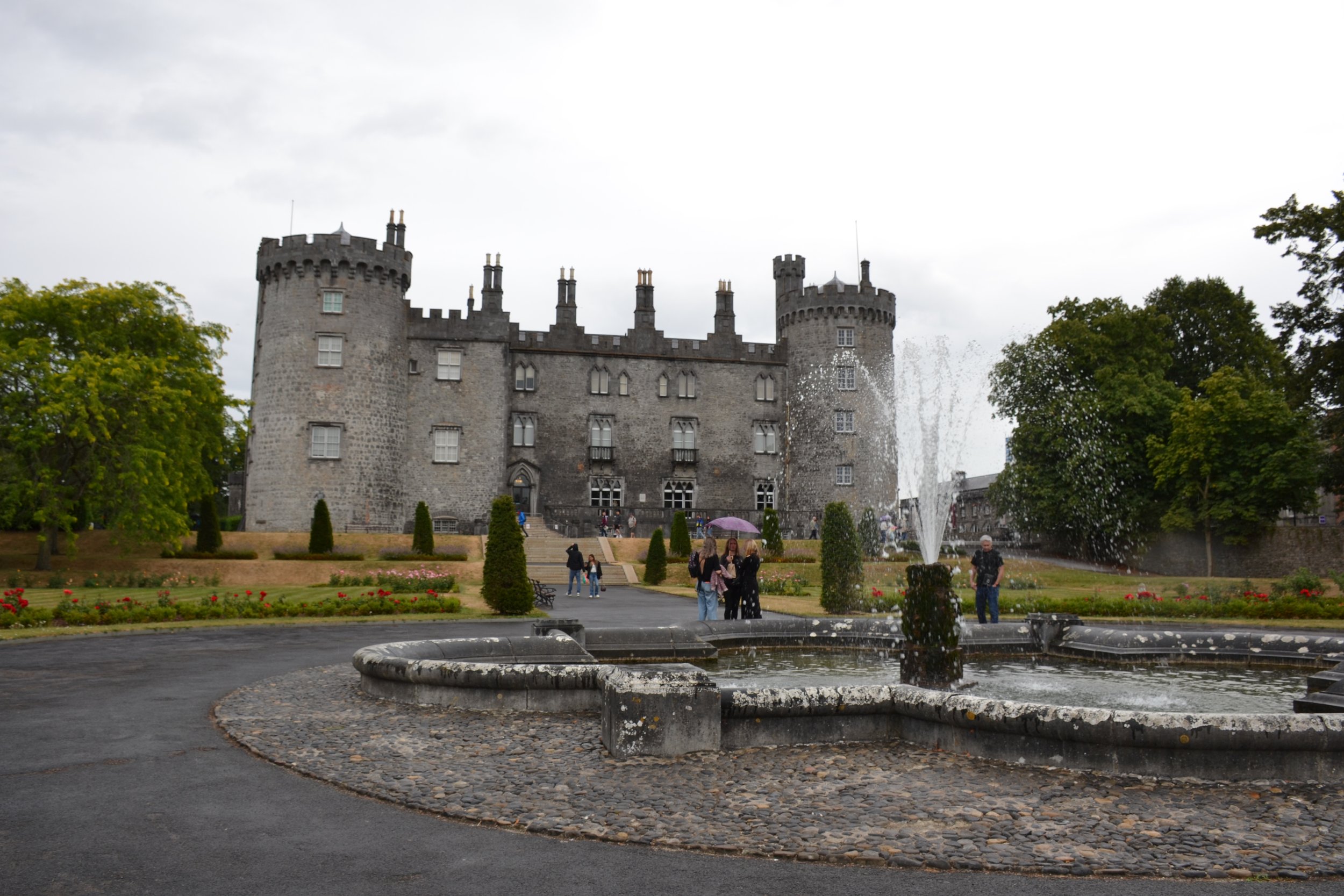 Overall view of Kilkenny Castle from the formal garden, showing its historic stone façade, round towers, crenellated roofline, landscaped grounds, and fountain.