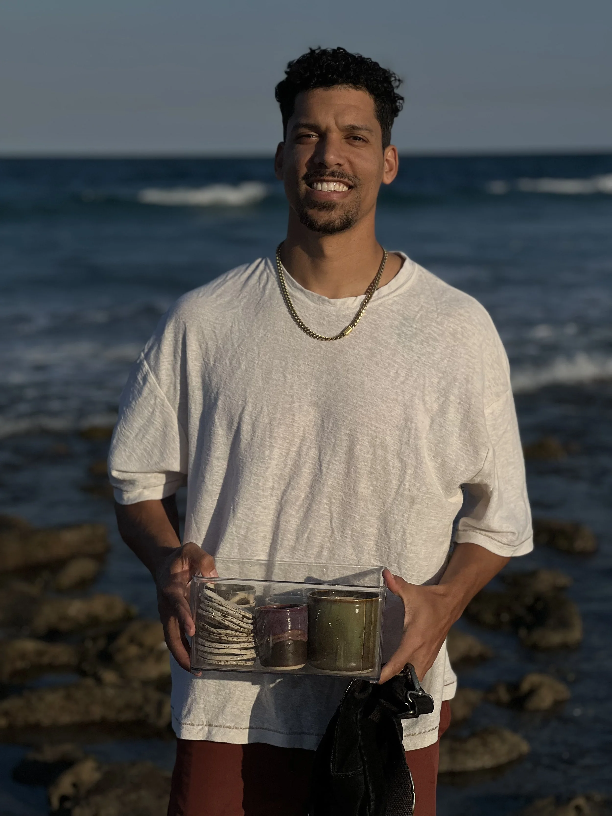 A man with curly black hair, a goatee, and wearing a light-colored t-shirt with a gold chain necklace, standing on a beach with rocks and the ocean in the background, holding a clear plastic container with three different colored cups or bowls inside.
