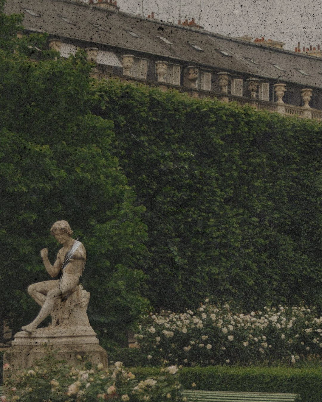 A garden with a classical statue of a seated woman on a pedestal, surrounded by lush green bushes and white flowers, with a building in the background.