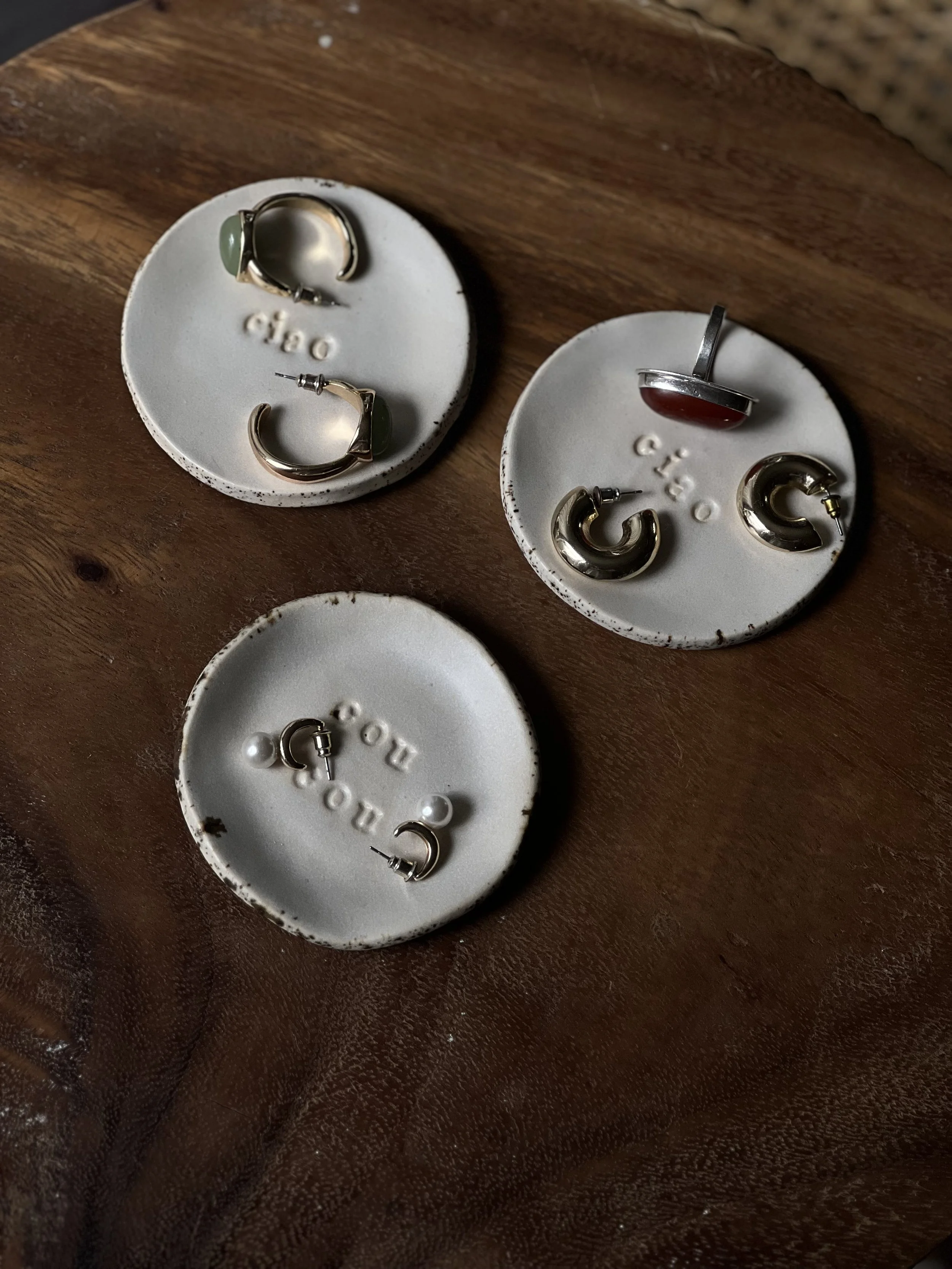 Three white ceramic jewelry display dishes on a wooden surface, each with earrings resting on them. The dishes have the word 'chao' embossed on their centers.