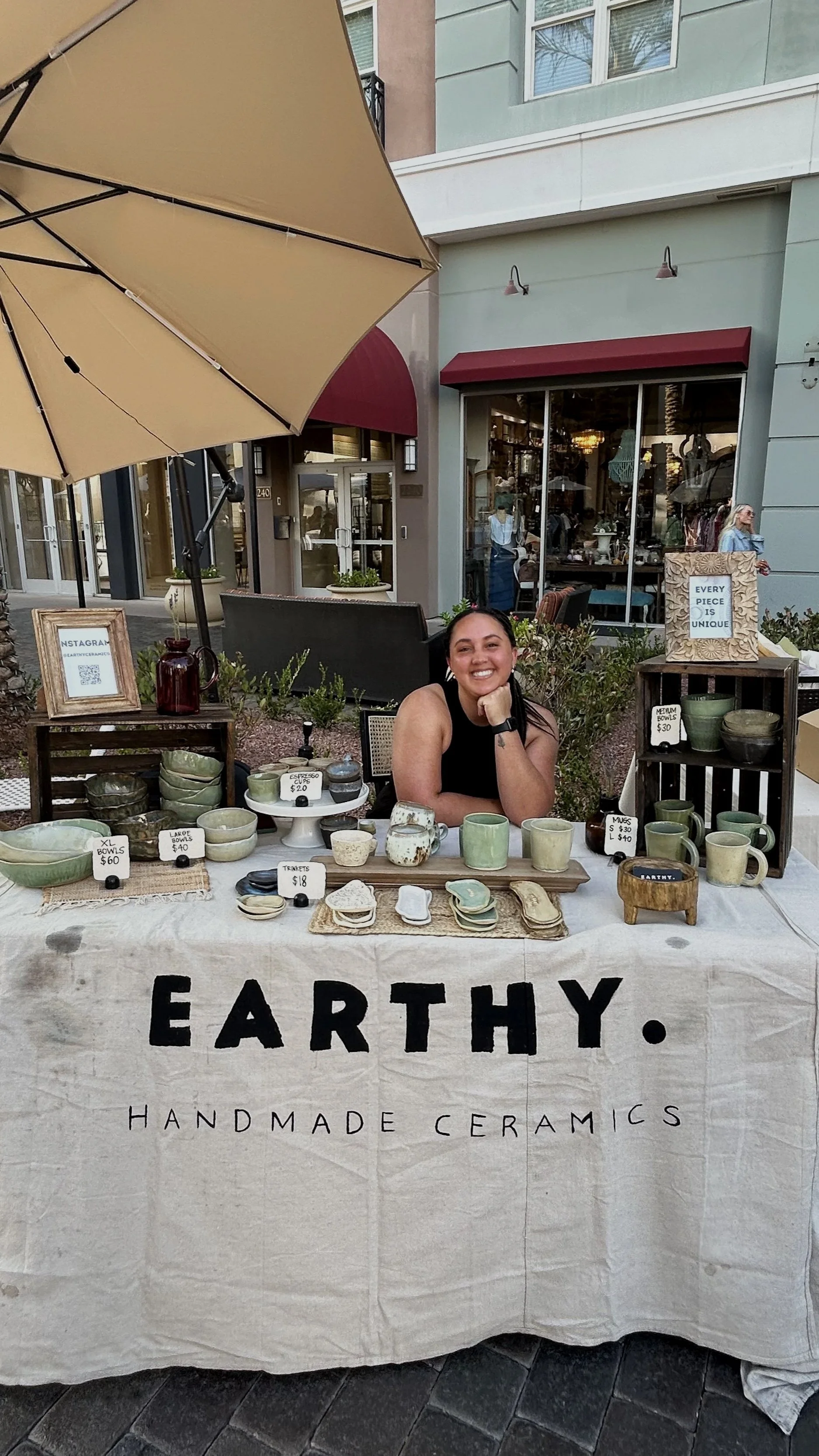 A woman sitting at a ceramic booth labeled 'EARTHY. HANDMADE CERAMICS' at a street market. The booth displays various handmade ceramic bowls, mugs, and trays for sale. There is a large beige umbrella providing shade, and behind the booth are storefronts and pedestrians.