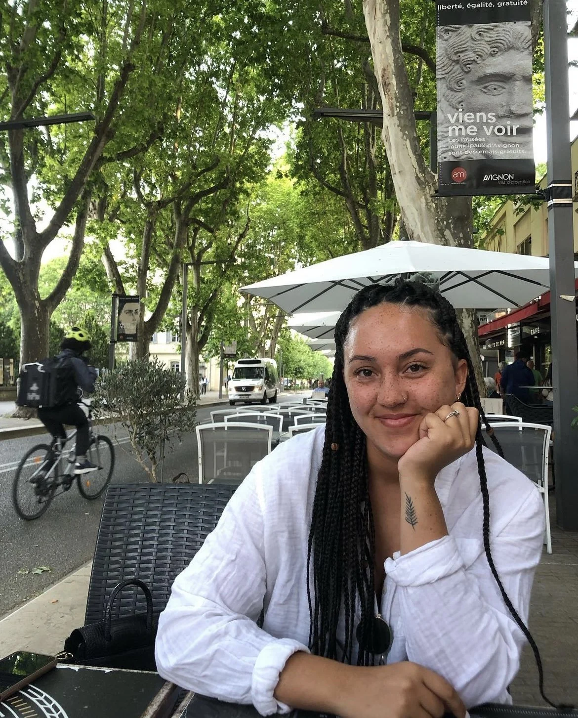 A young woman with braided hair and a tattoo on her hand sitting at an outdoor cafe, smiling at the camera, with trees and a cyclist passing by in the background.