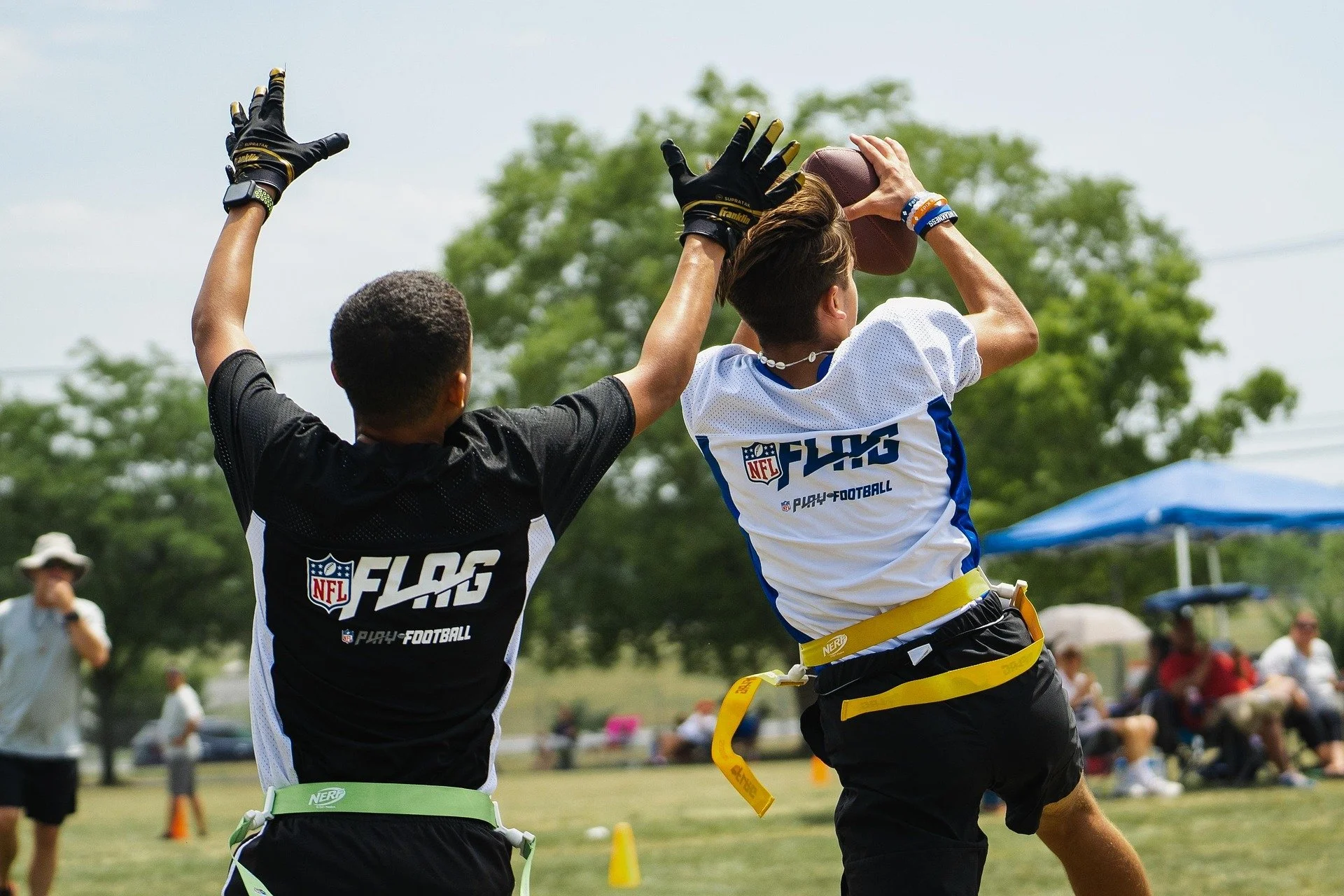 Two young football players wearing NFL FLAG shirts competing in a flag football game outdoors, one is attempting to catch a football while the other is trying to block.