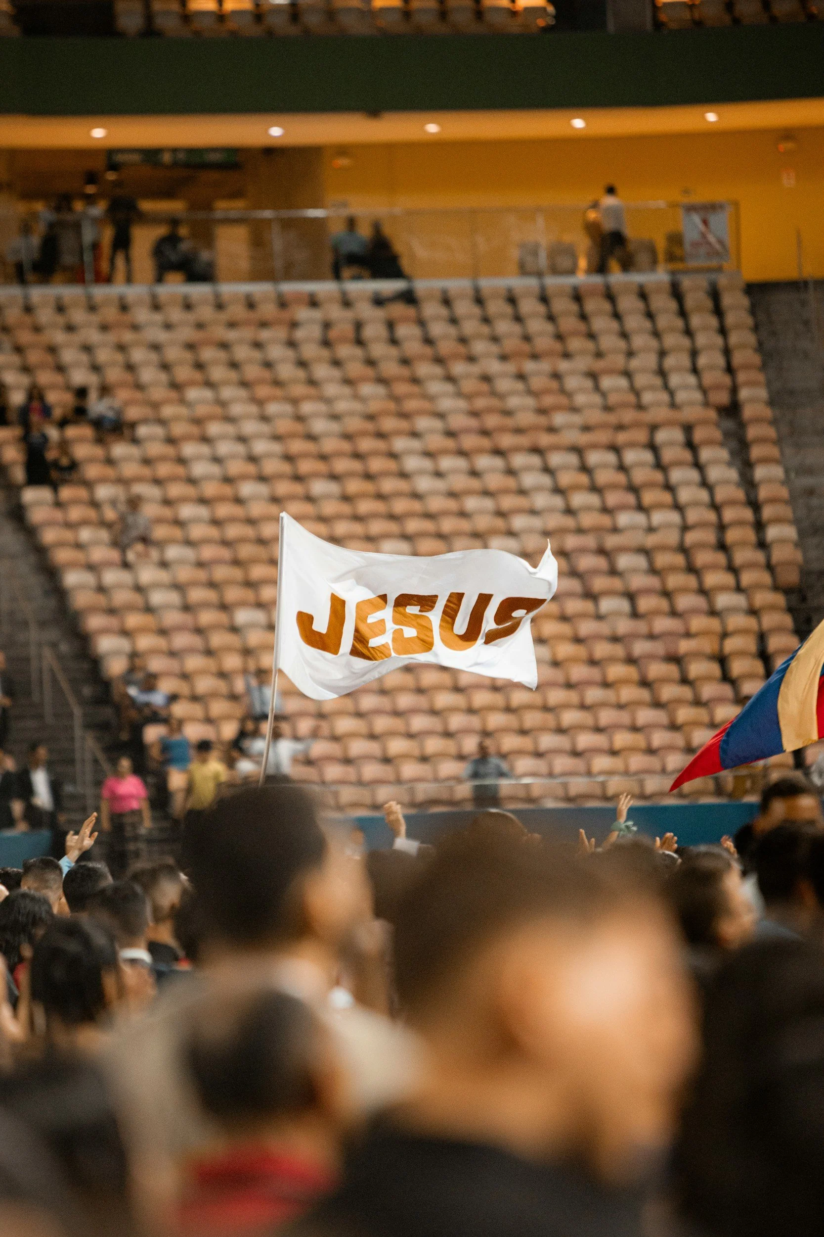 Crowd of people at a gathering or event in an indoor stadium or auditorium, with a flag reading 'JESUS' being waved amid others in the audience.