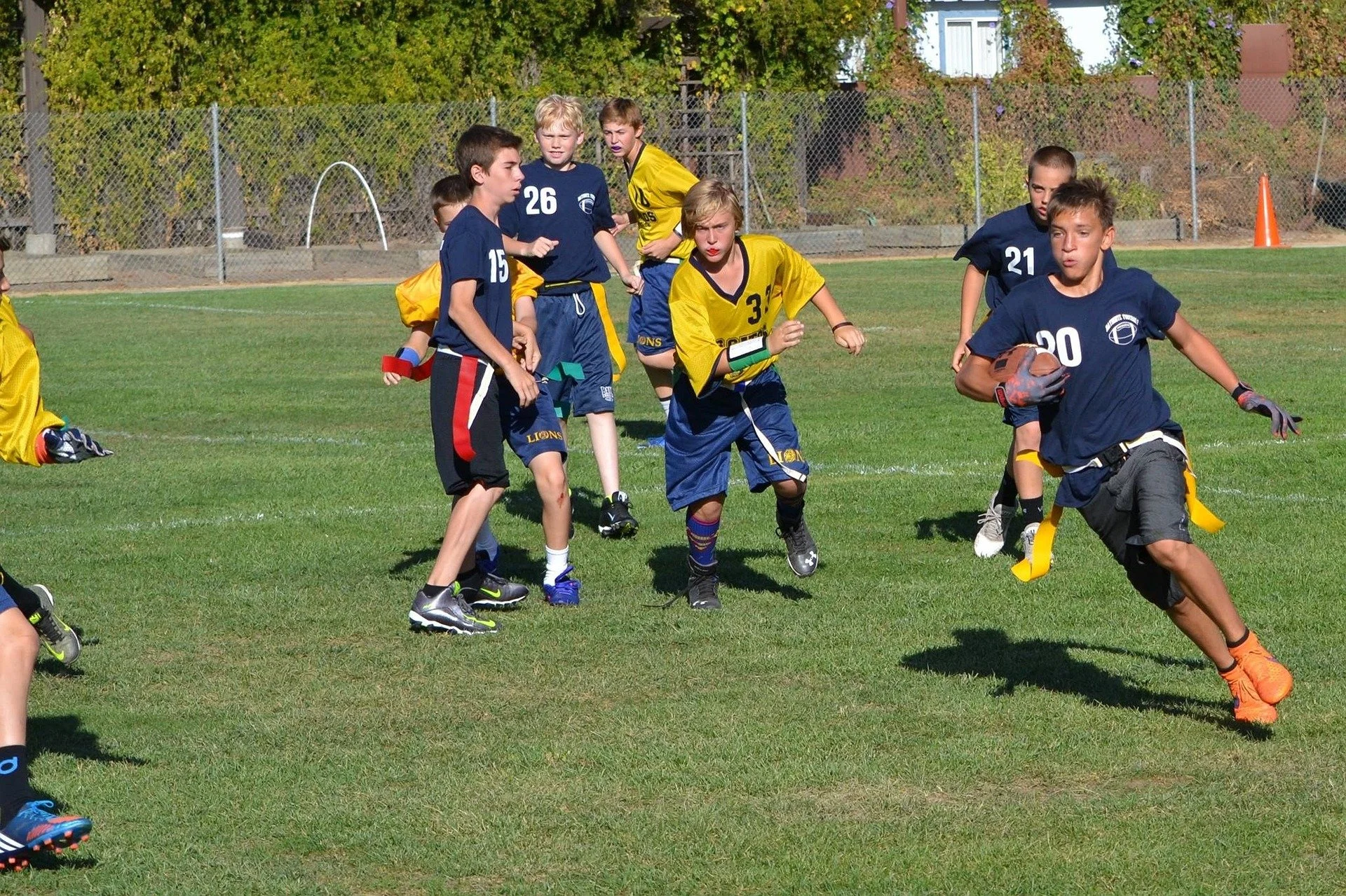 Young boys playing flag football on a grassy field during daytime, some wearing blue jerseys and others in yellow, with one boy running with a football.