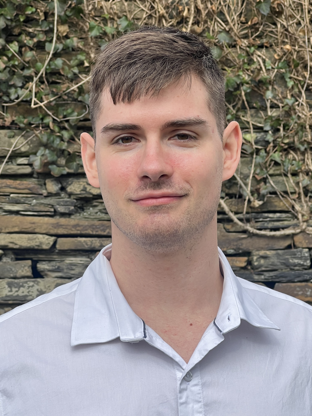Portrait of a young man with short brown hair, wearing a white collared shirt, standing outdoors in front of a stone wall with green ivy.
