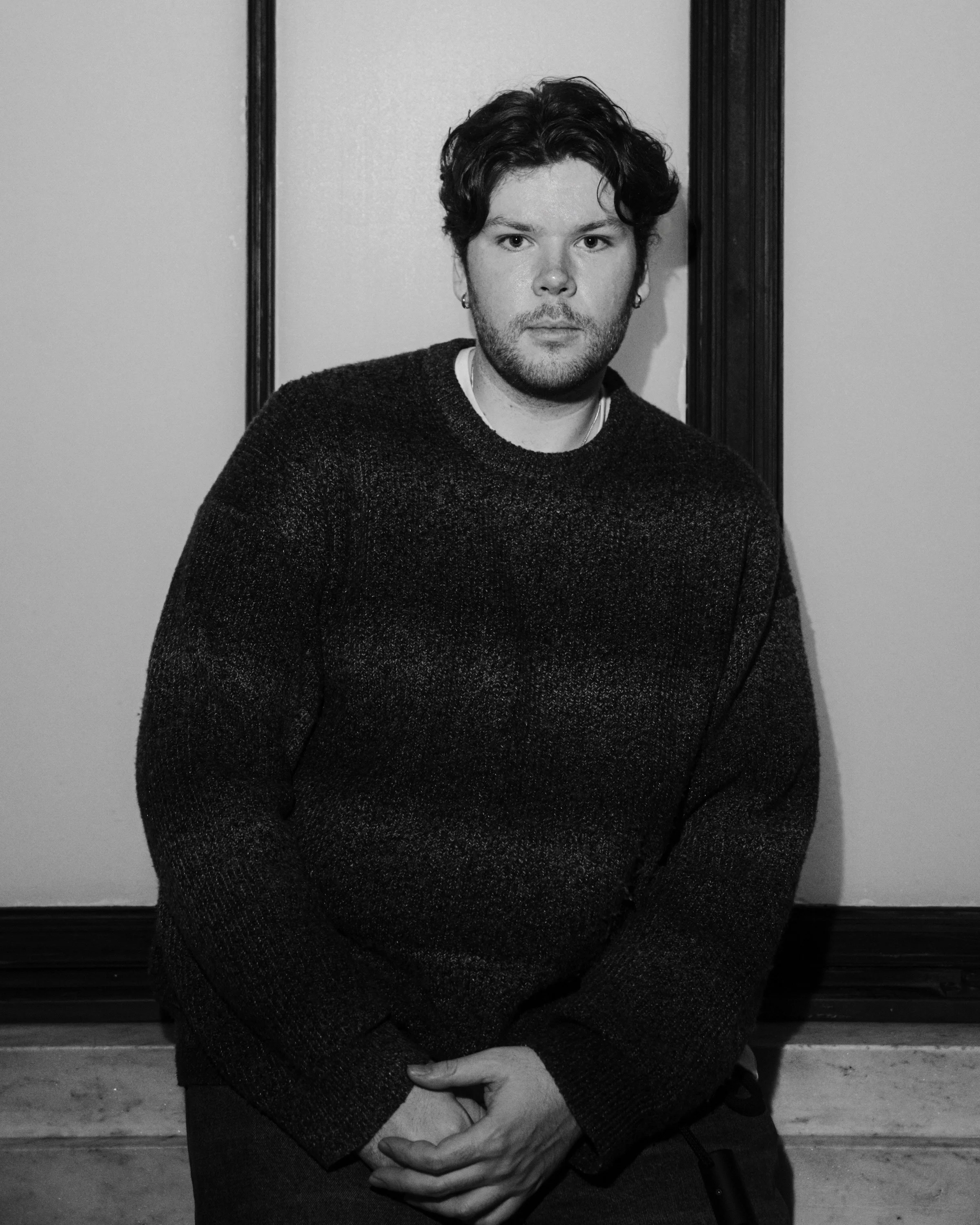 A black and white photo of a young man with curly hair, wearing a dark sweater, sitting and looking directly at the camera. He is in front of a wall with dark trim.