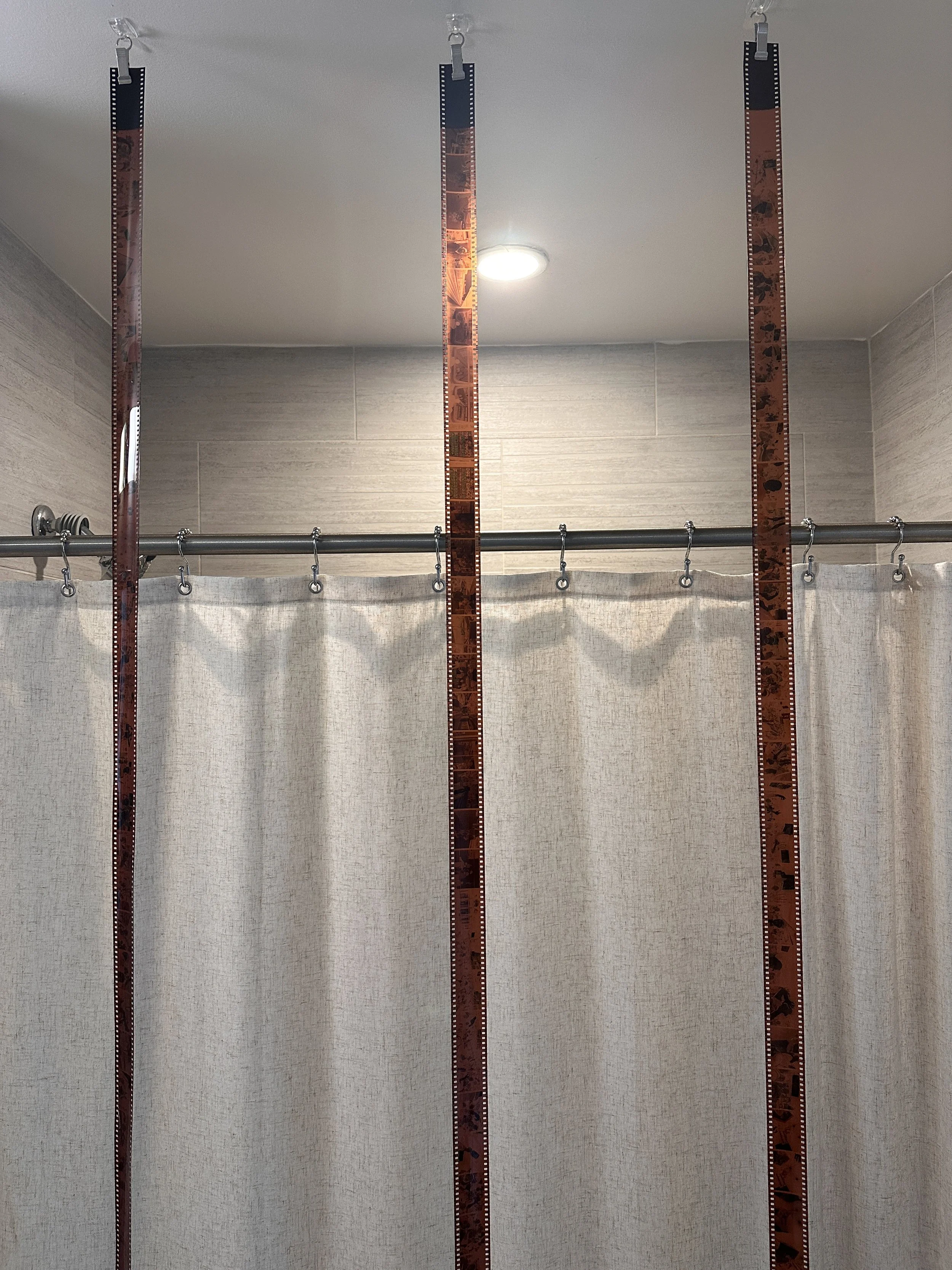 Three vertical measuring tapes hanging from hooks, positioned in front of a shower curtain with a metal rod and two hooks, inside a bathroom with tiled walls and a ceiling light.