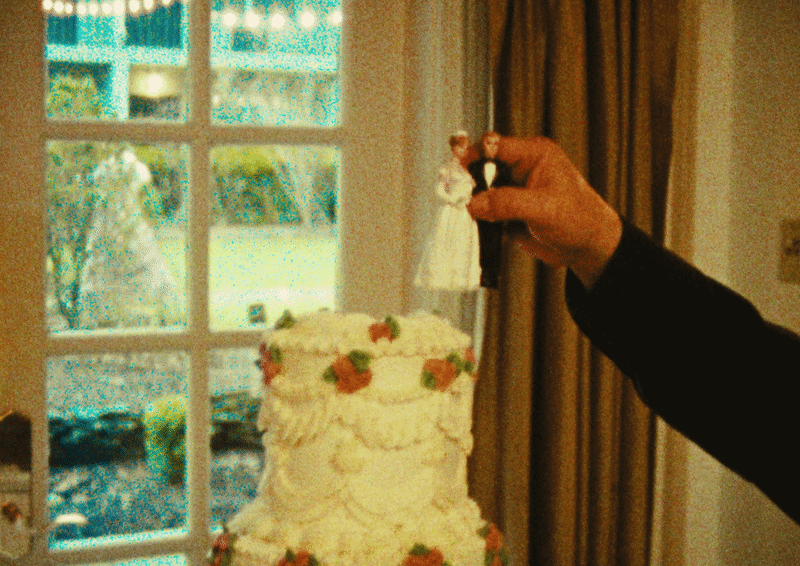 A person's hand holding a bride and groom figurine above a wedding cake with flowers, in front of a window with curtains.