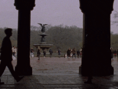 View through an archway of a Betheseda Terrace with a fountain and statue, surrounded by people, with trees and overcast sky in the background in Central Park.