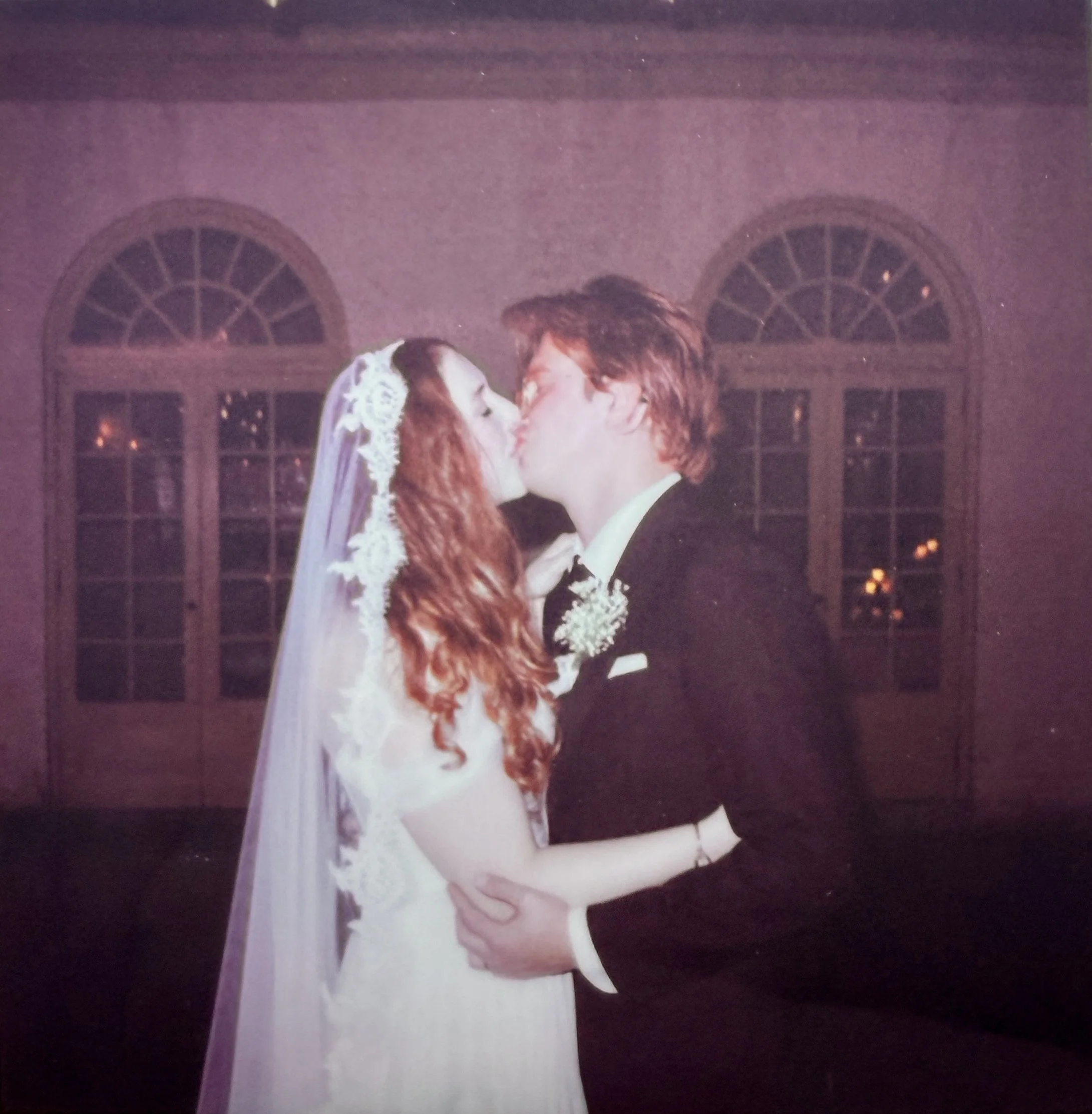 A bride and groom sharing a kiss on their wedding day in an indoor venue with arched windows in the background.