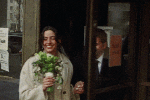 A woman is holding a bouquet of flowers and smiling as she walks out of a building with a man following her.