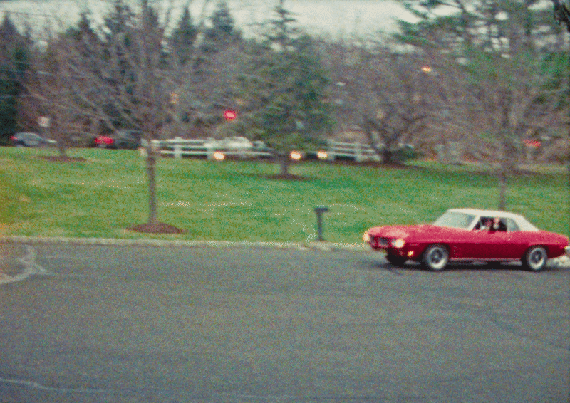 A red vintage car parked on a paved area near a grassy park with trees and a small mailbox.