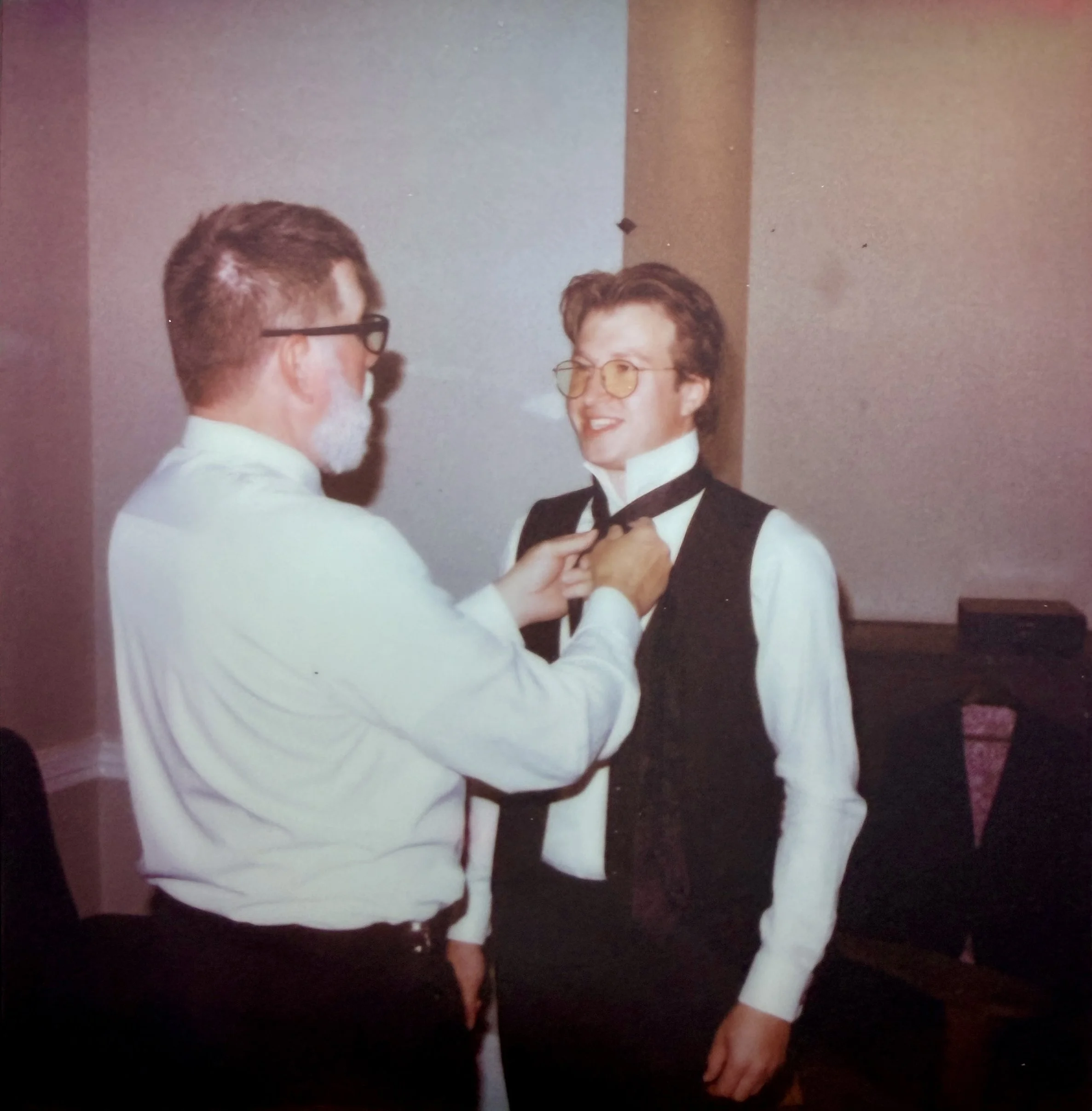 A young man in a tuxedo cosplaying as Elton John with glasses and a bow tie is being helped with his bow tie by an older man with glasses, a gray beard, and a white shirt. They are inside a room with a plain wall background.