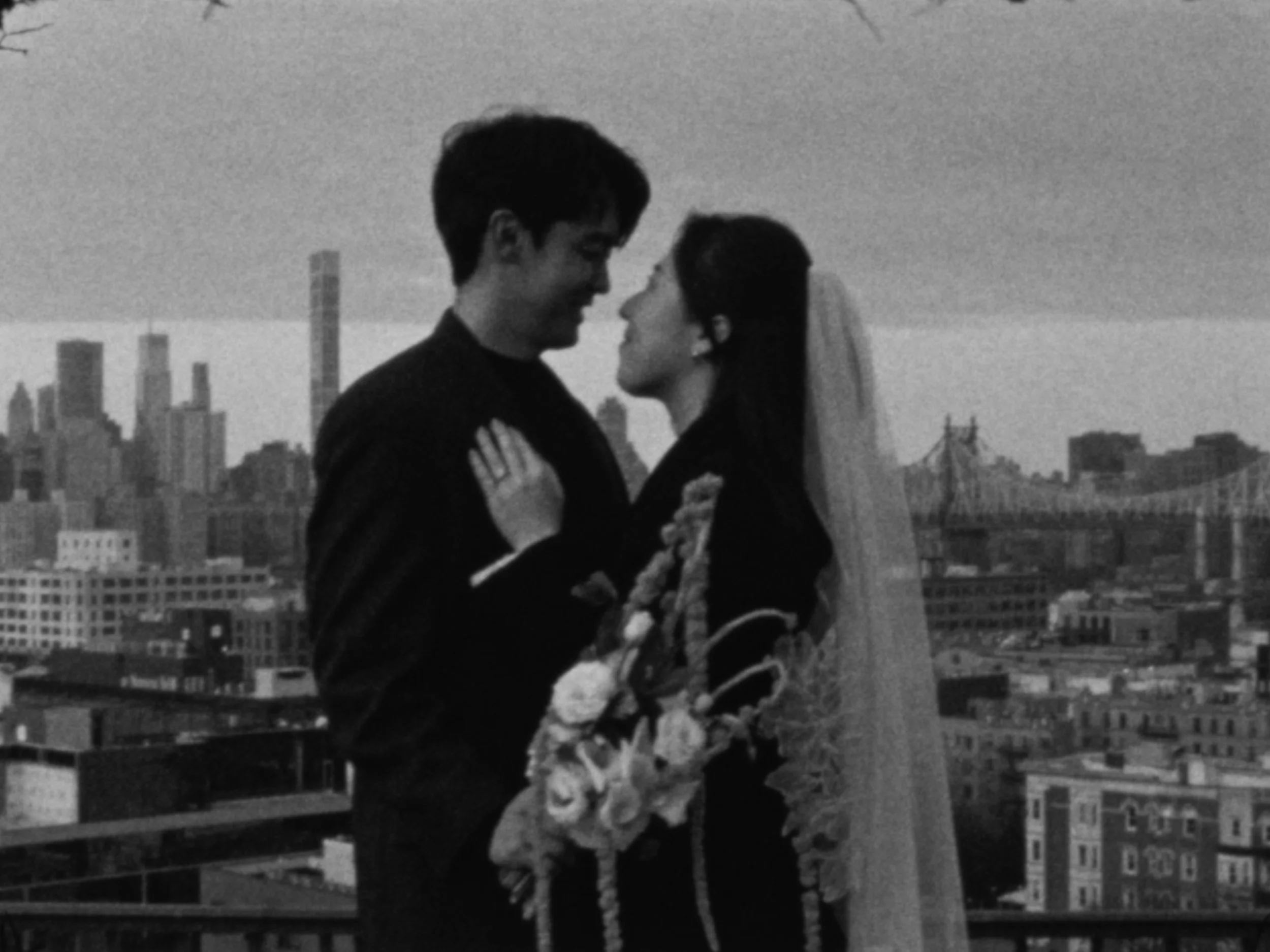 A smiling young man and woman embracing on a rooftop with a city skyline and a bridge in the background.