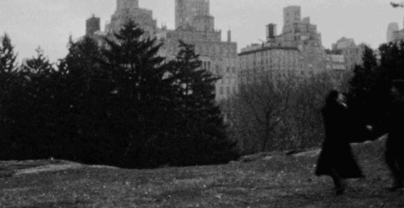 Black and white photo of two people walking in a park with city buildings in the background.
