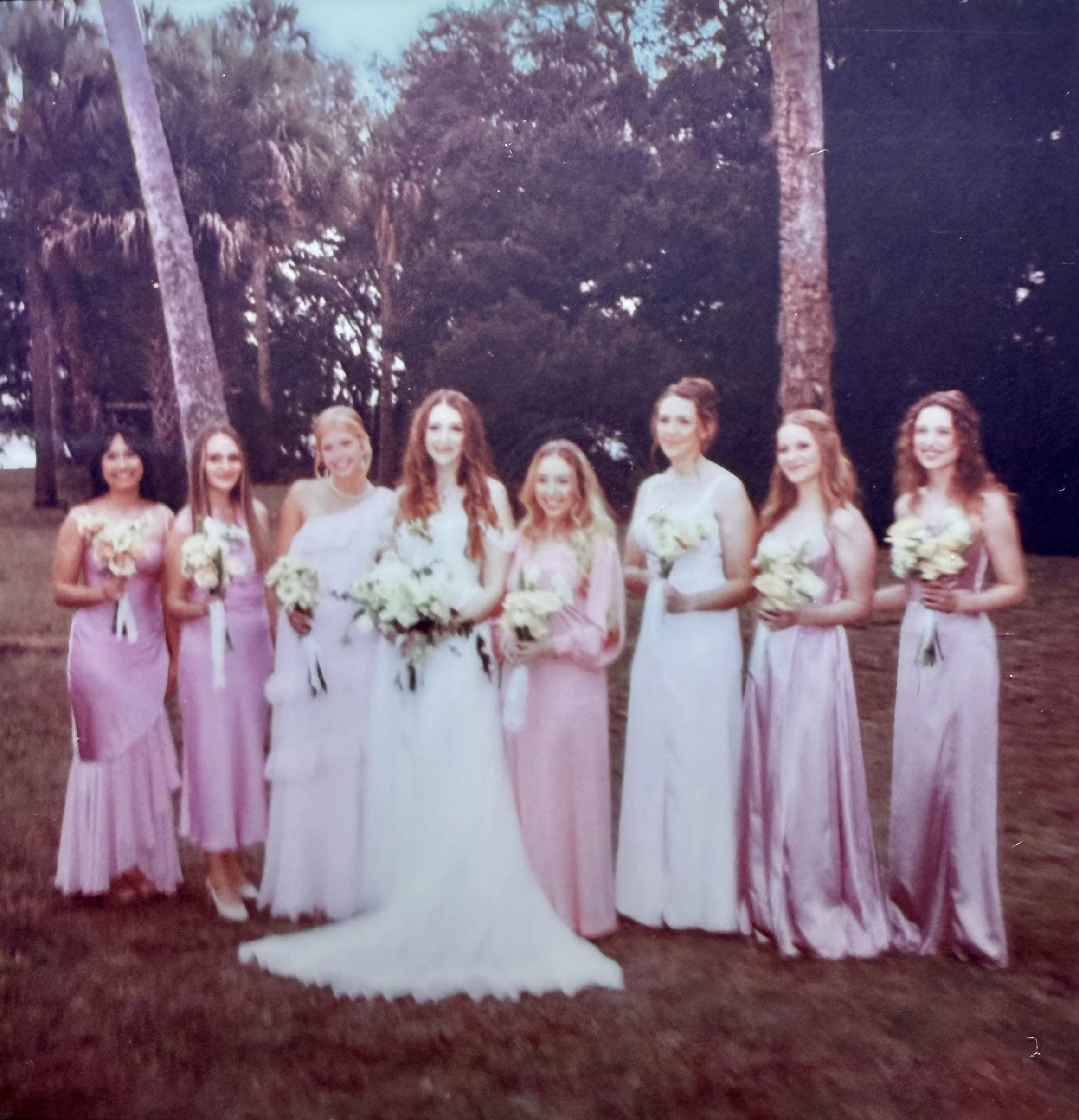 A group of eight women, dressed in pink and white dresses, standing outdoors on grass with tall trees in the background, holding bouquets of flowers.