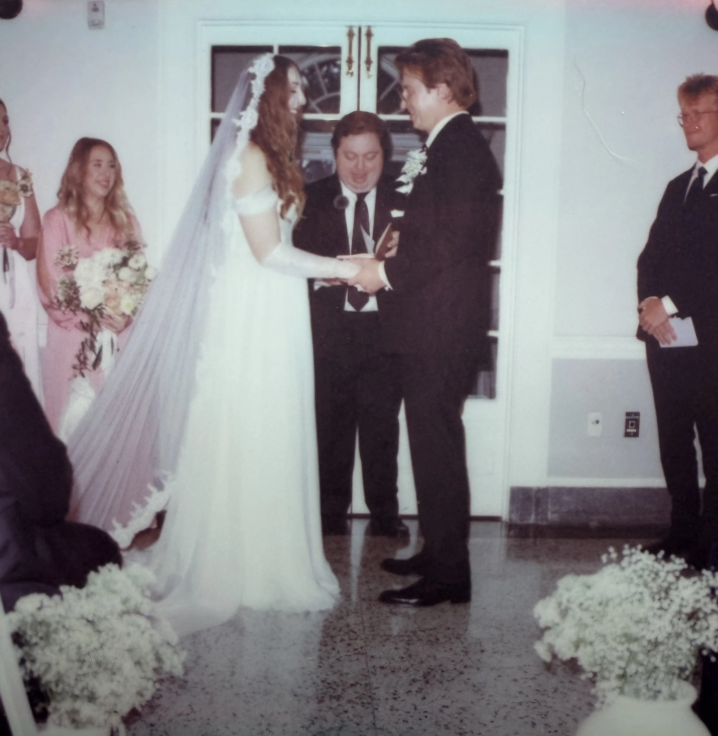 A bride and groom exchanging vows during a wedding ceremony indoors, with bridesmaids and groomsmen standing nearby.