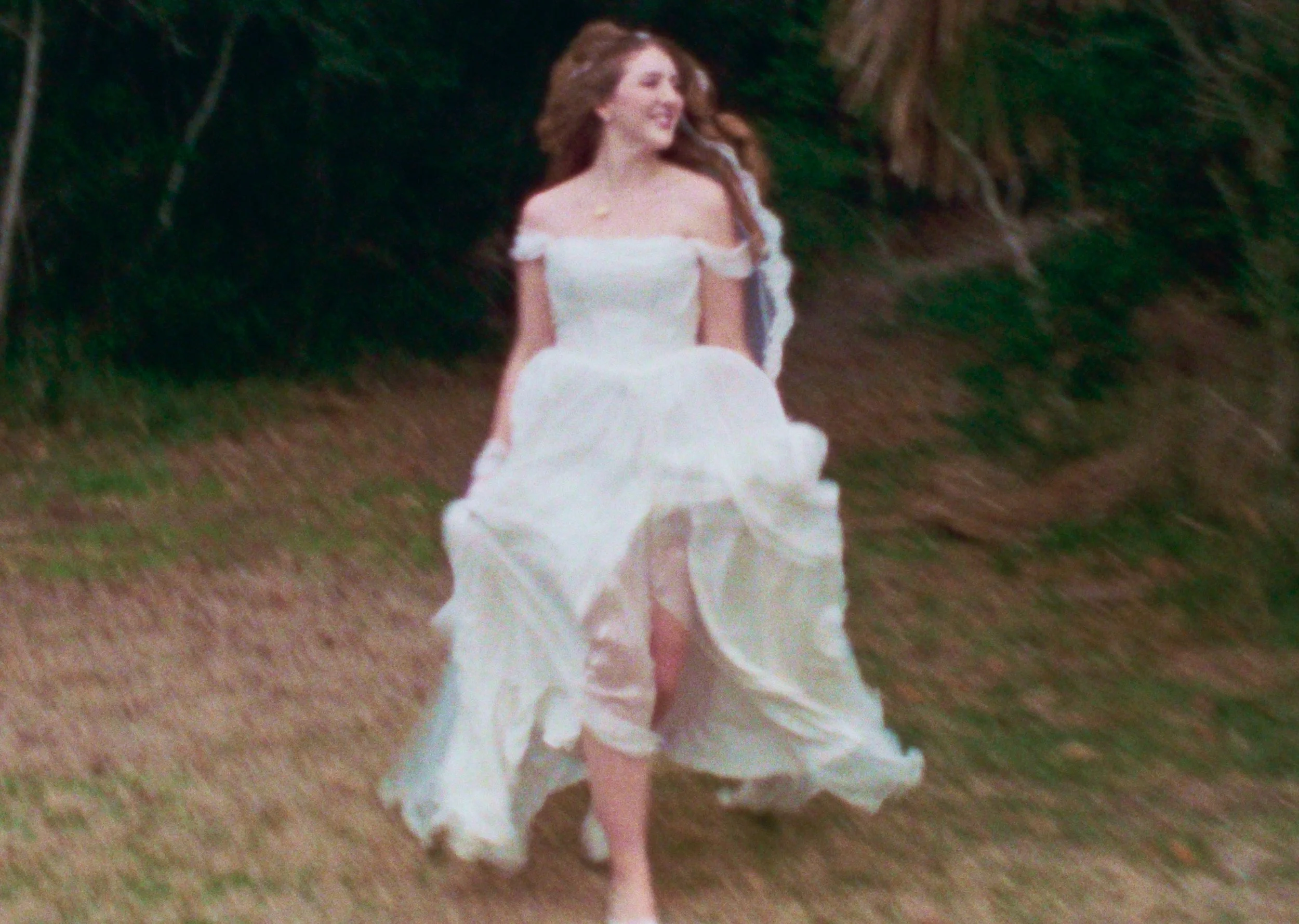 A woman in a white off-shoulder dress walking outdoors, with a wooded background.