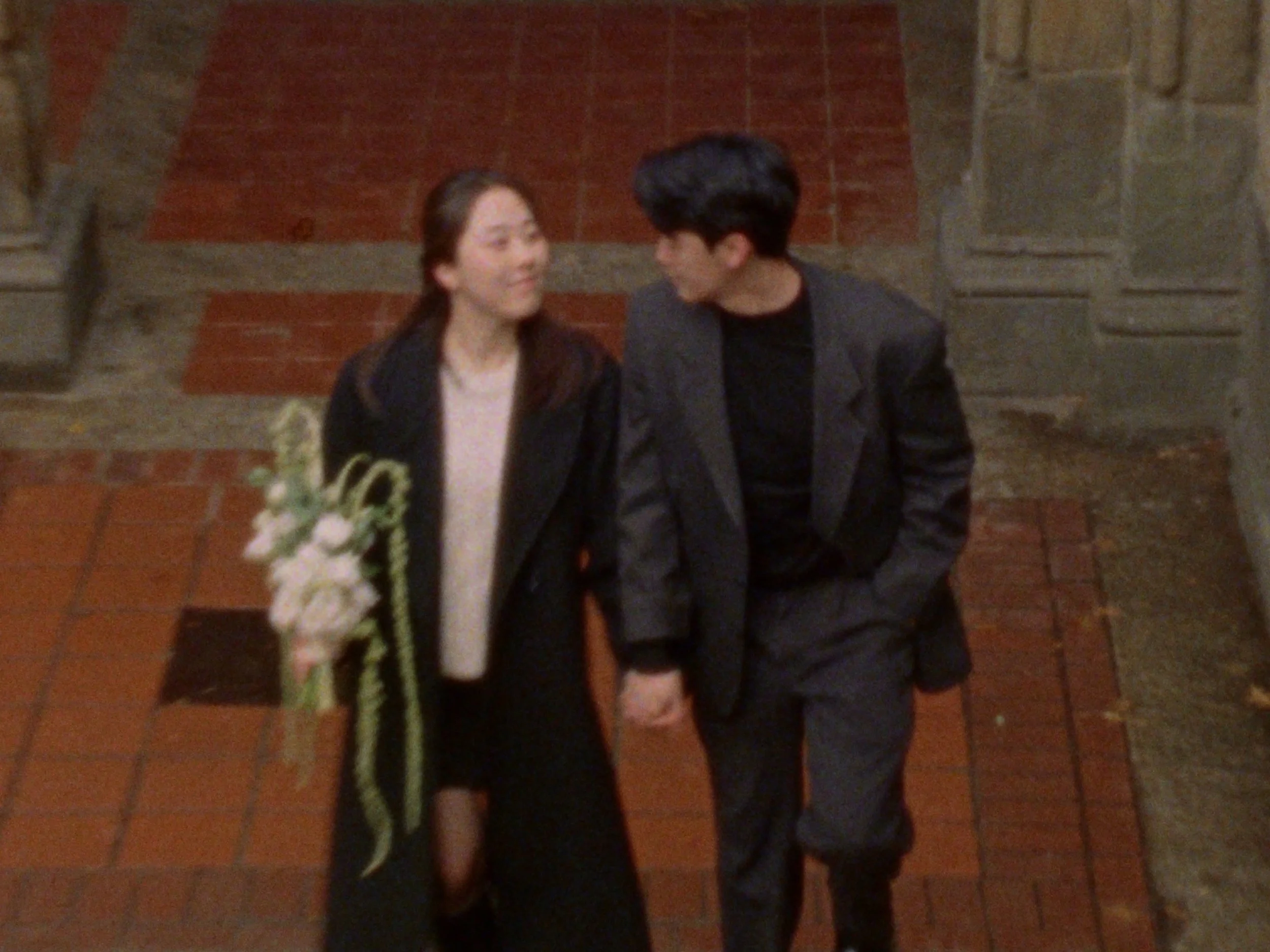 A couple dressed in formal attire holding a bouquet of flowers, standing close together on a railway track during evening or nighttime with benches and autumn-colored trees in the background.