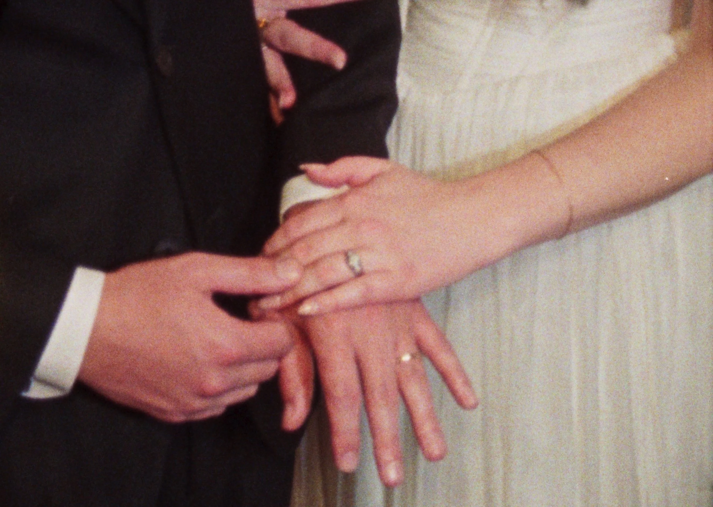 A person in a black suit is holding the hand of a person in a white dress, showing off rings on both their ring fingers, likely during a wedding ceremony.