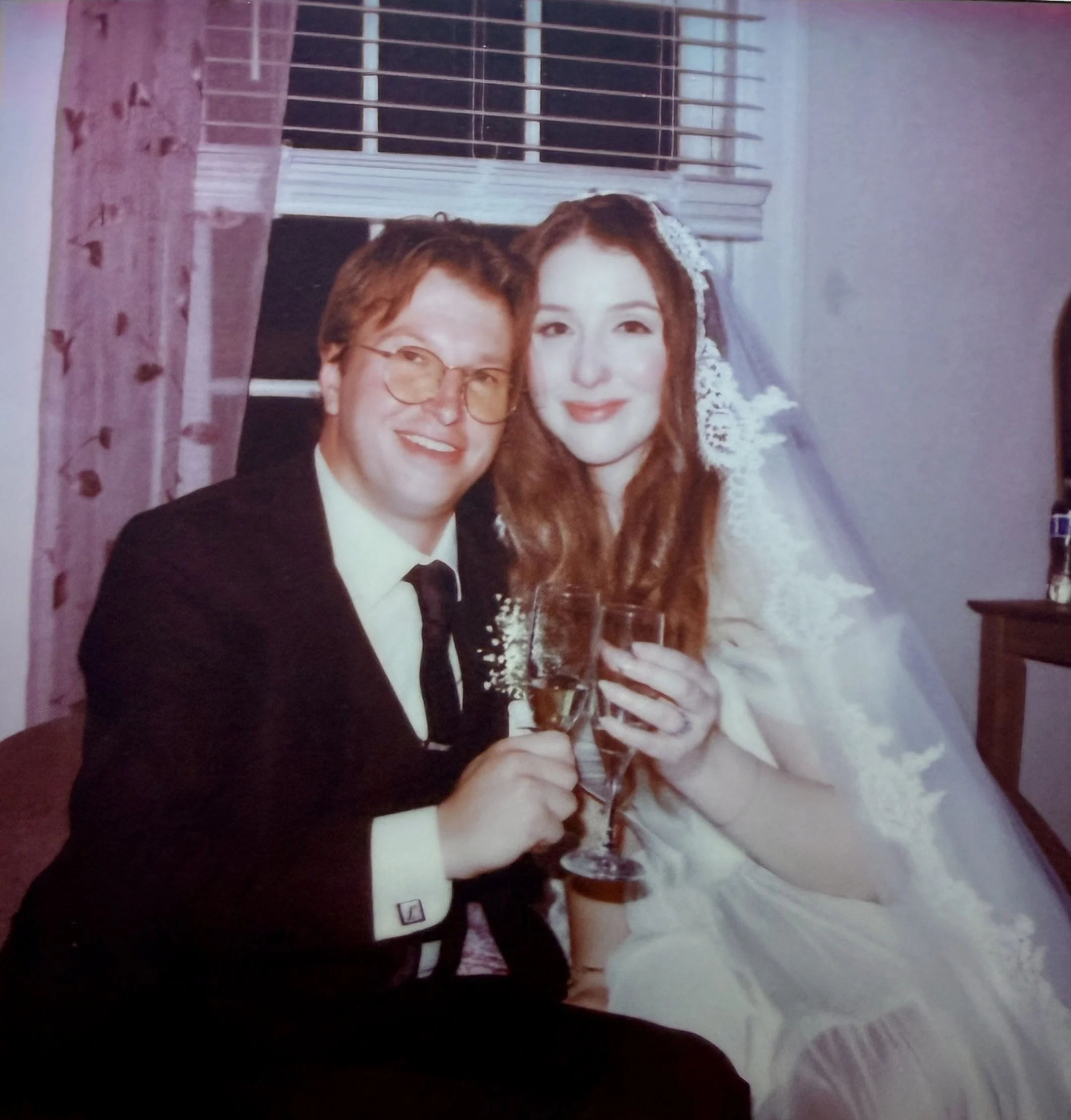 A man in a tuxedo and a woman in a wedding dress holding glasses of champagne pose for a photo together. They are smiling and appear to be celebrating a wedding.
