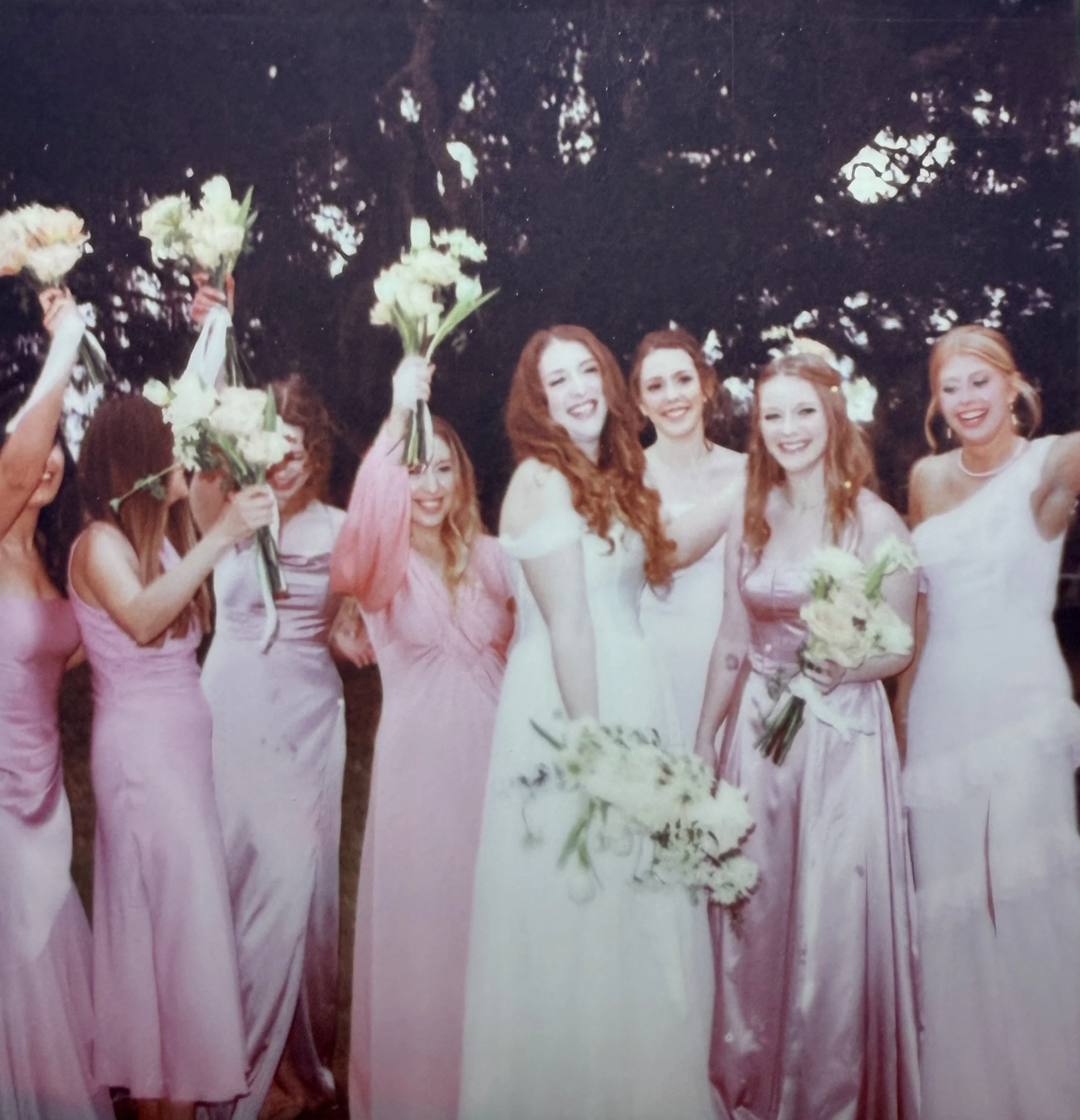 Group of women at a wedding, with bride in white gown and bridesmaids in pink dresses, holding bouquets of white flowers outside during evening.