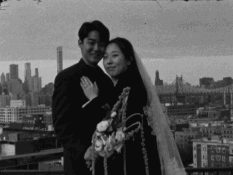 A black-and-white photo of a bride and groom smiling on a rooftop with a city skyline in the background, holding a bouquet of flowers.