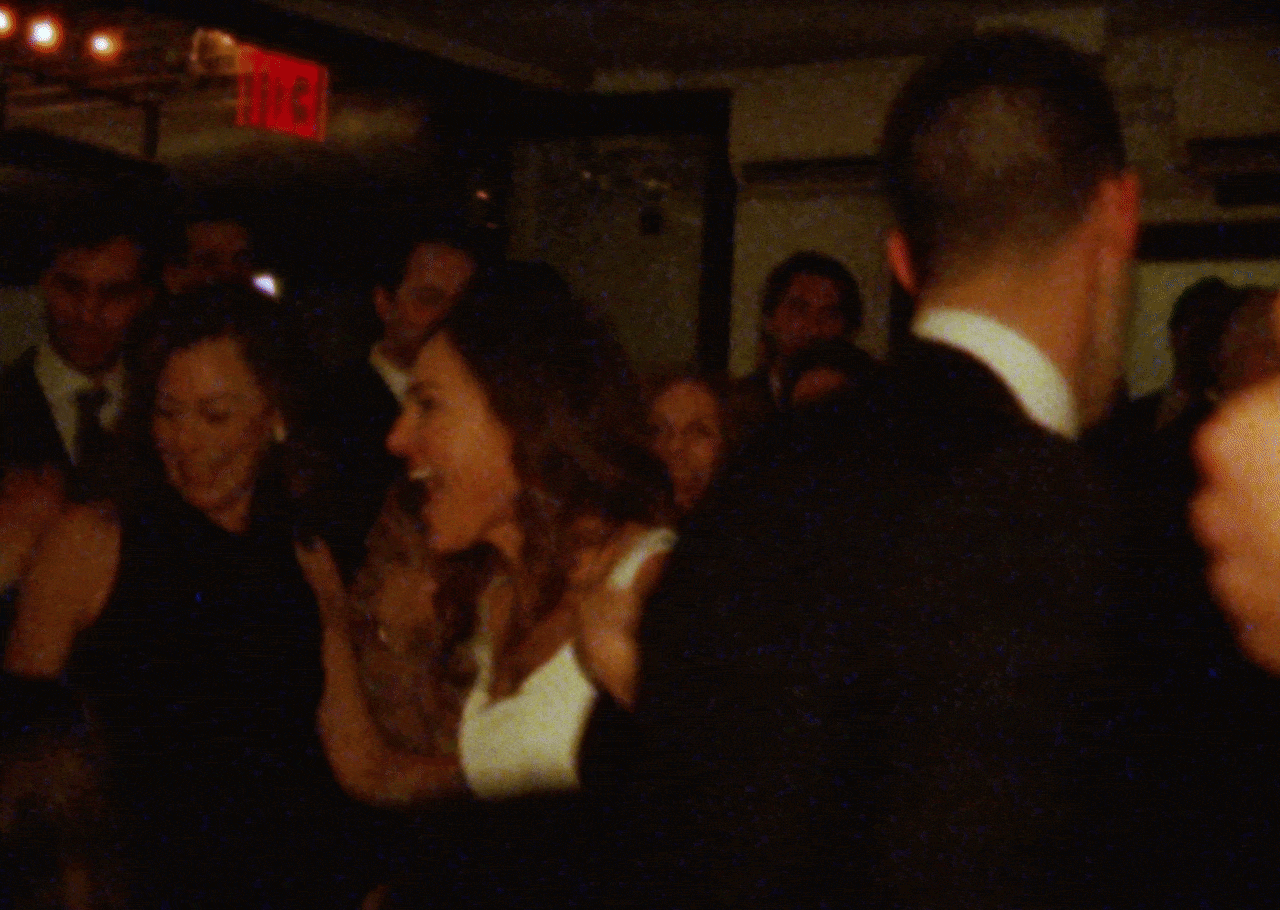 Bride & groom celebrating and dancing at a wedding, dressed in formal attire with a dark background and warm lighting.