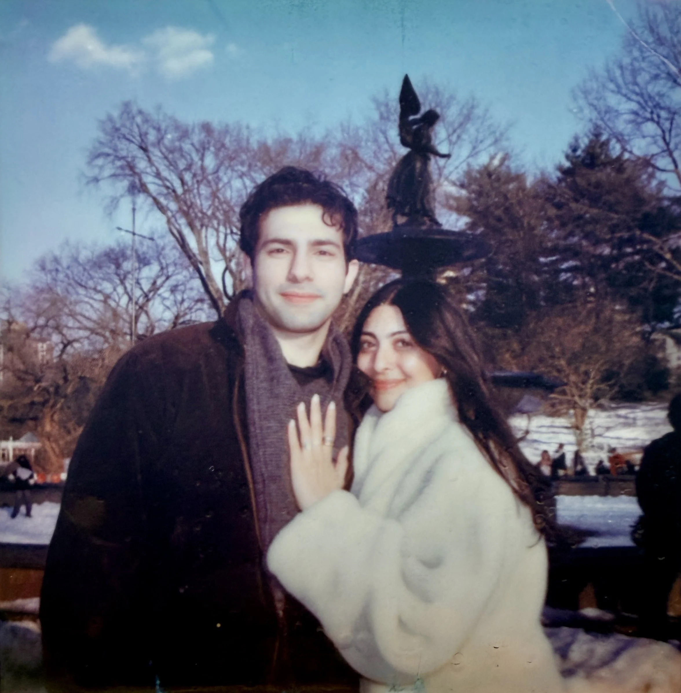 A young couple with light skin in winter clothing posing outdoors in front of a leafless tree and a snow-covered park, with the woman wearing a white coat and a ring on her finger, and the man wearing a dark jacket and scarf. The woman is balancing a small witch figurine on her head.