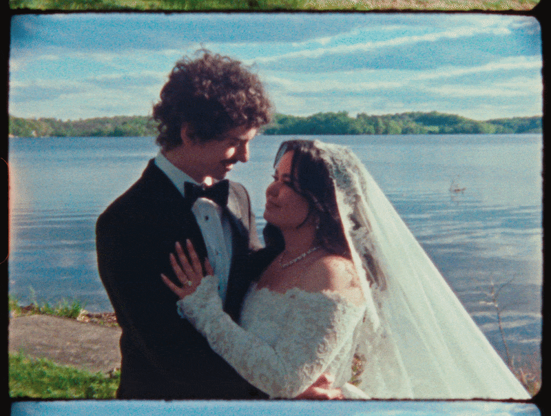 A bride and groom in wedding attire standing close together near a body of water, with trees in the background and a partly cloudy sky.