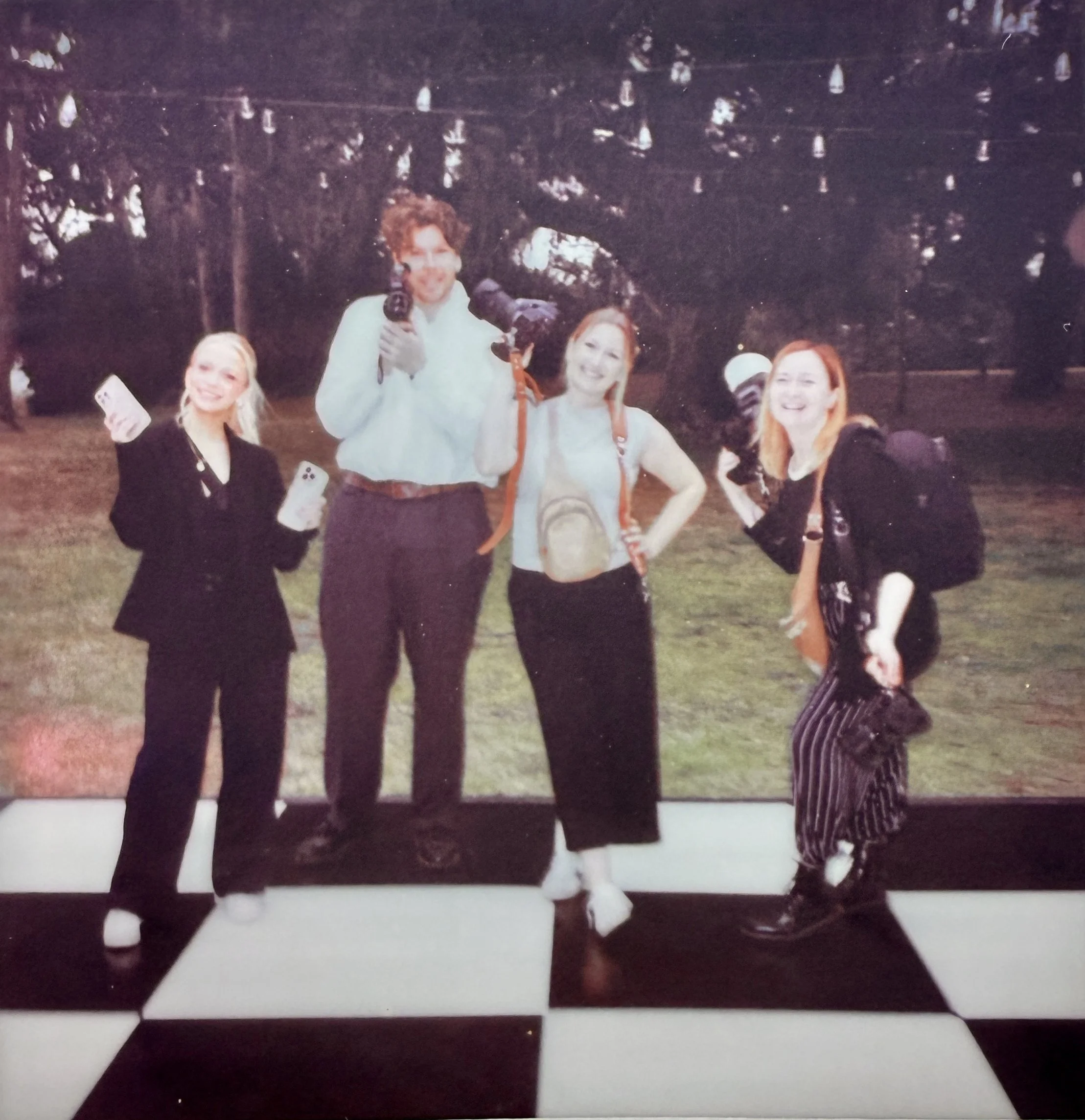 Four people standing outdoors on a black and white checkered floor, smiling and holding phones or cameras, with trees in the background.