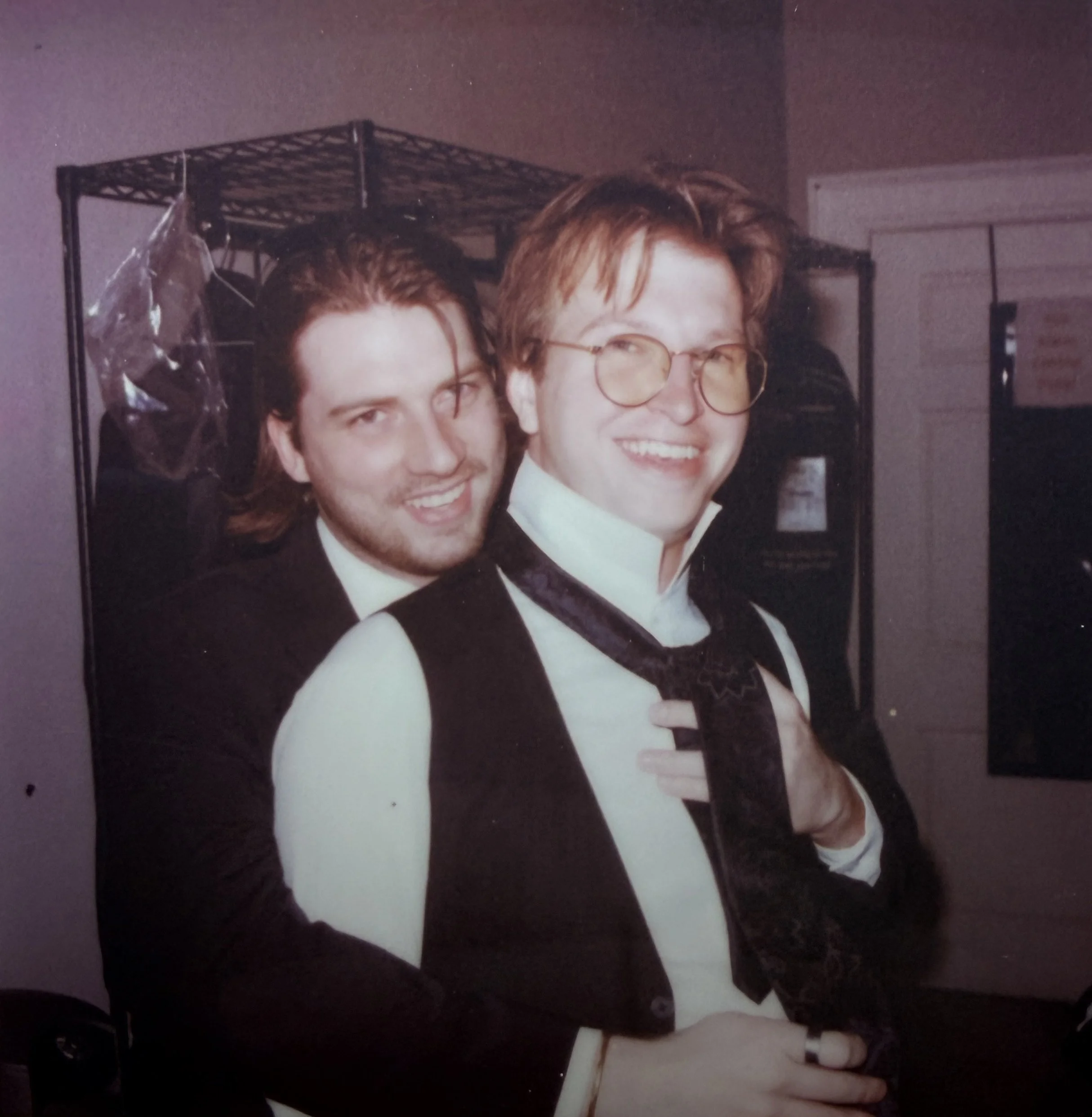 Two men smiling, one with long hair and beard, wearing a tuxedo with a white shirt, the other with red hair and glasses, wearing a black vest with a white shirt and a black scarf, in a room with a metal shelf in the background.