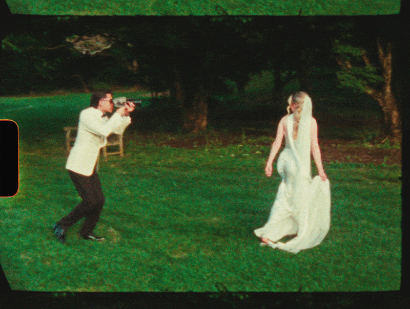 A man is taking a photograph of a woman in a white dress outdoors on a grassy area, with trees in the background.