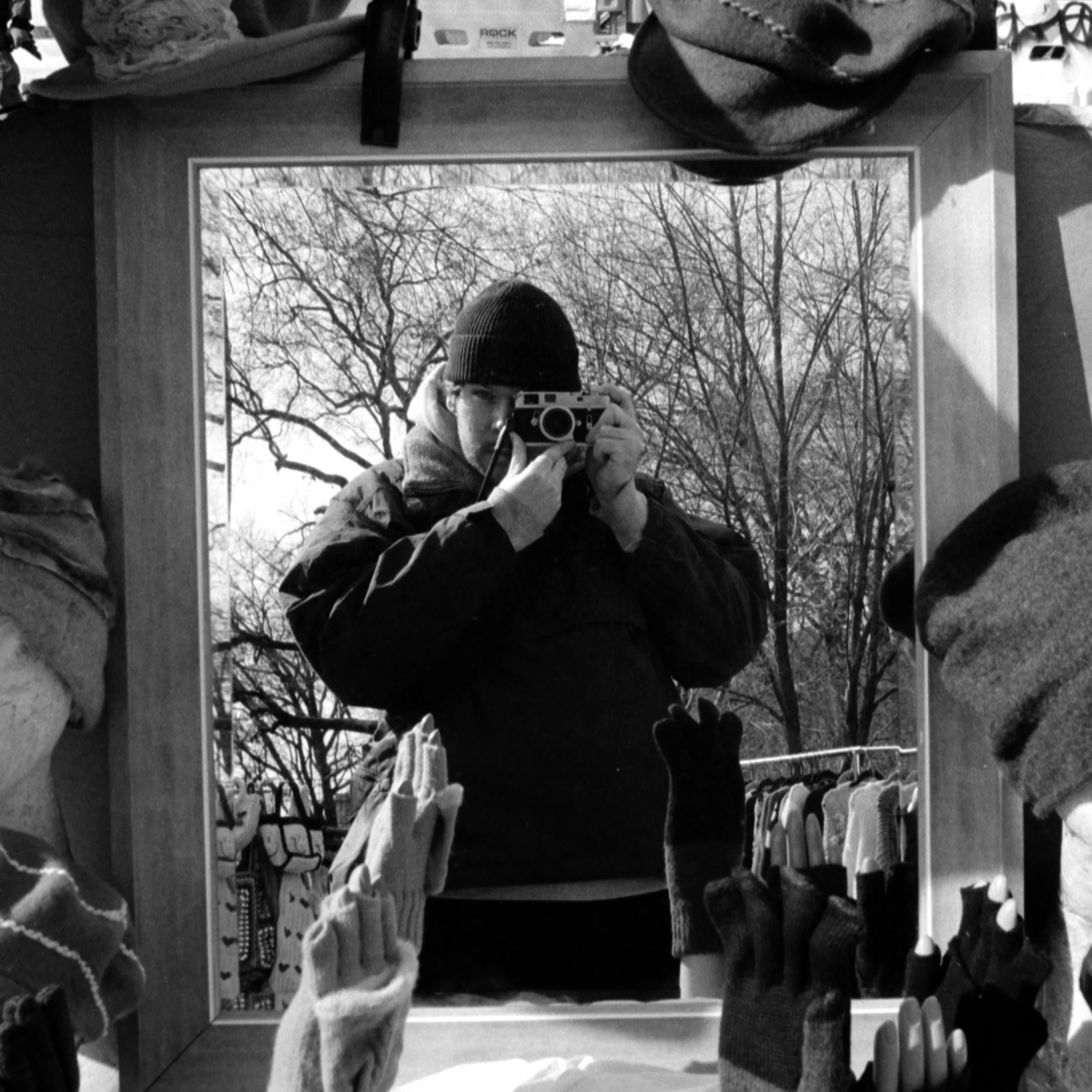 A person taking a photo of their reflection in a mirror at a thrift store, surrounded by clothing and accessories, with trees visible outside.