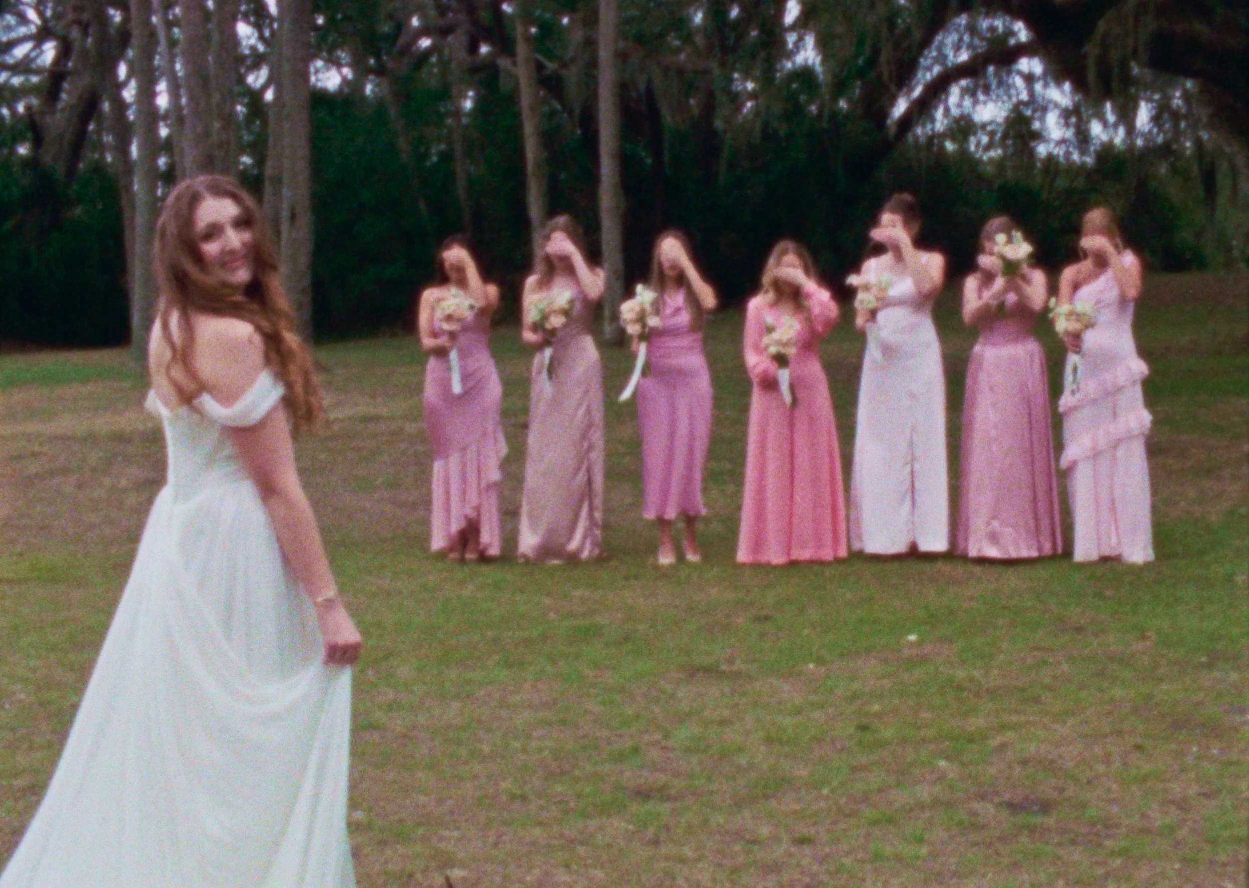 A bride in a white wedding gown looking back and smiling, with seven bridesmaids in pink and white dresses in the background holding bouquets, standing outdoors on a grassy area surrounded by trees.