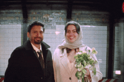 A man and a woman standing together indoors, the woman holding a bouquet of white and purple flowers, both smiling. Infront of the New York City subway, during a courthouse wedding.