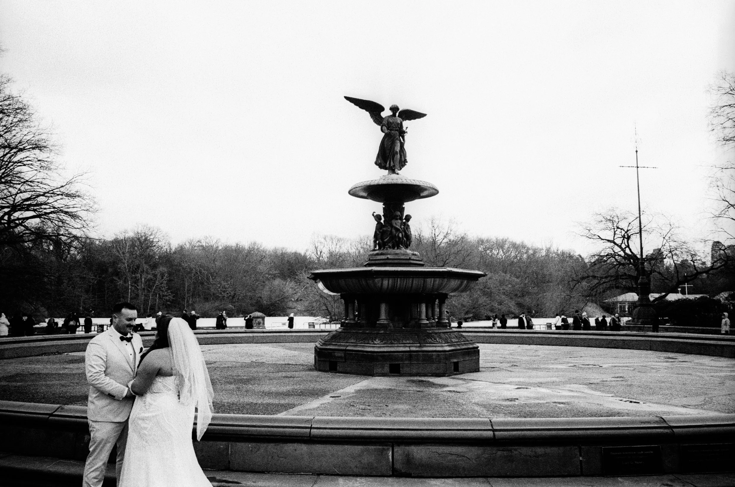A bride and groom dressed in wedding attire, holding hands, standing in front of a large fountain with a statue of an angel with wings on top. The scene is outdoors in a park with trees and a cloudy sky.