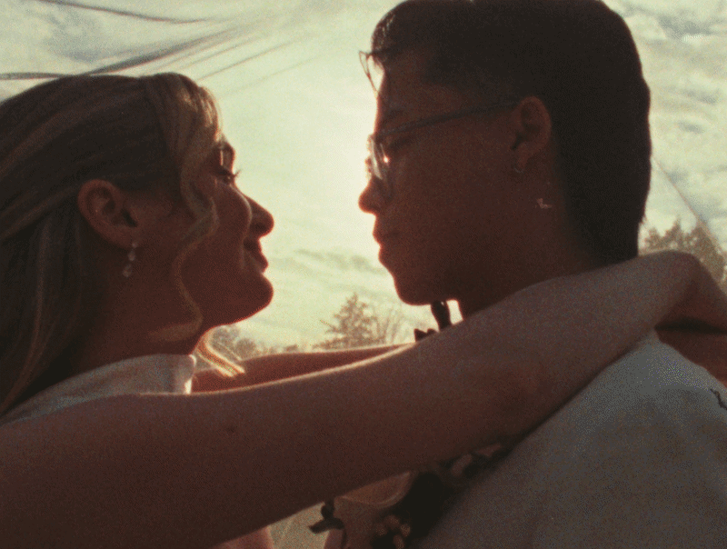 A woman and a man are close, looking into each other's eyes with the sunset behind them, outdoors.