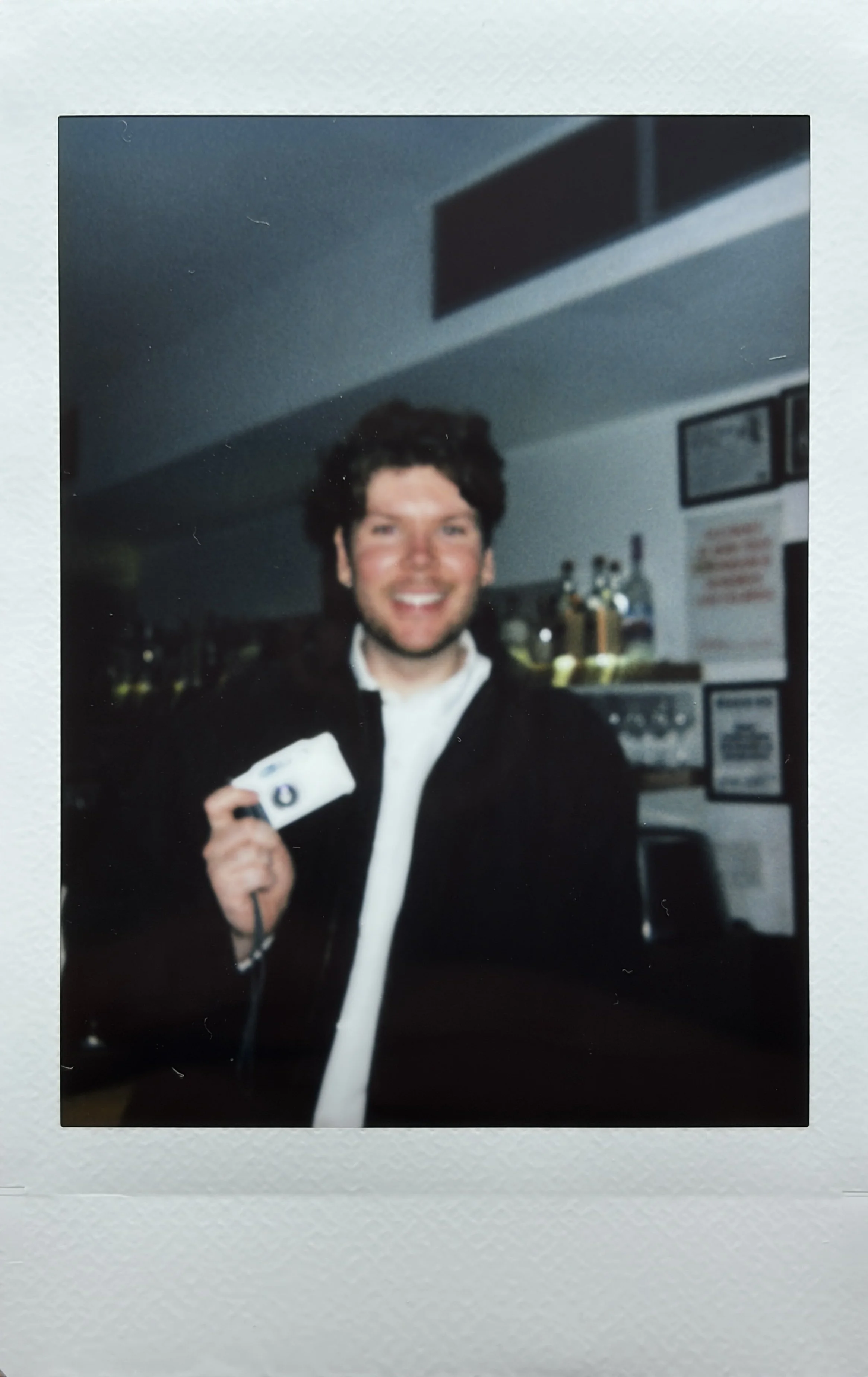 A smiling man with dark, curly hair holding a small device or camera, standing in front of a bar or counter with bottles and framed signs or menus on the wall behind him.