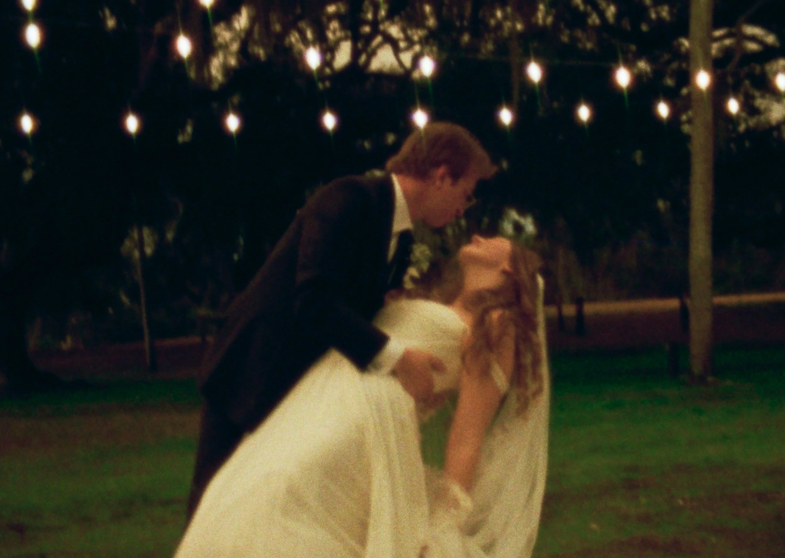 A couple dancing outdoors at night under string lights, with the man wearing a tuxedo and the woman in a wedding dress.