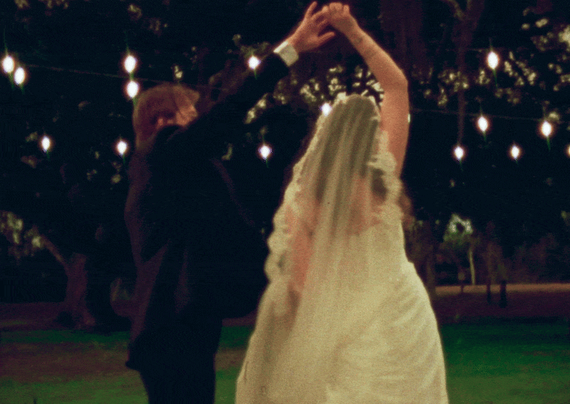 A couple dancing outdoors at night under string lights, with the woman in a white dress and the man in a dark suit.
