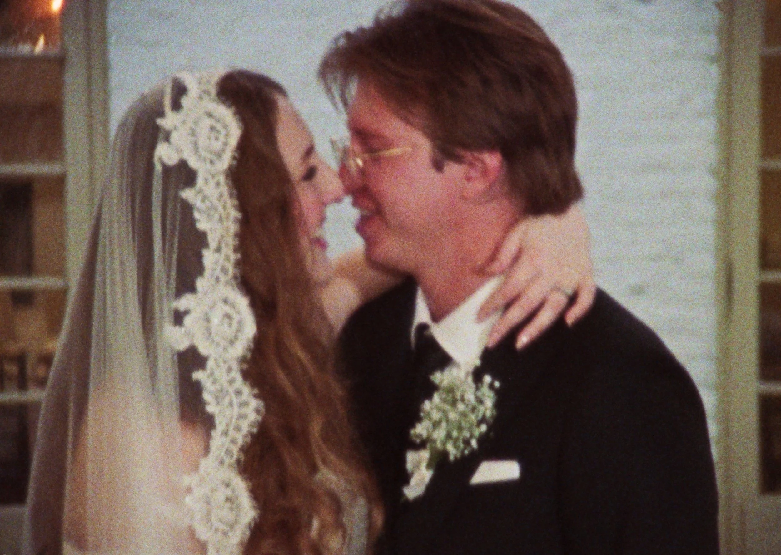 A close-up of a bride and groom touching foreheads and smiling at each other during their wedding. The bride has long hair and a lace veil, and the groom is wearing glasses.