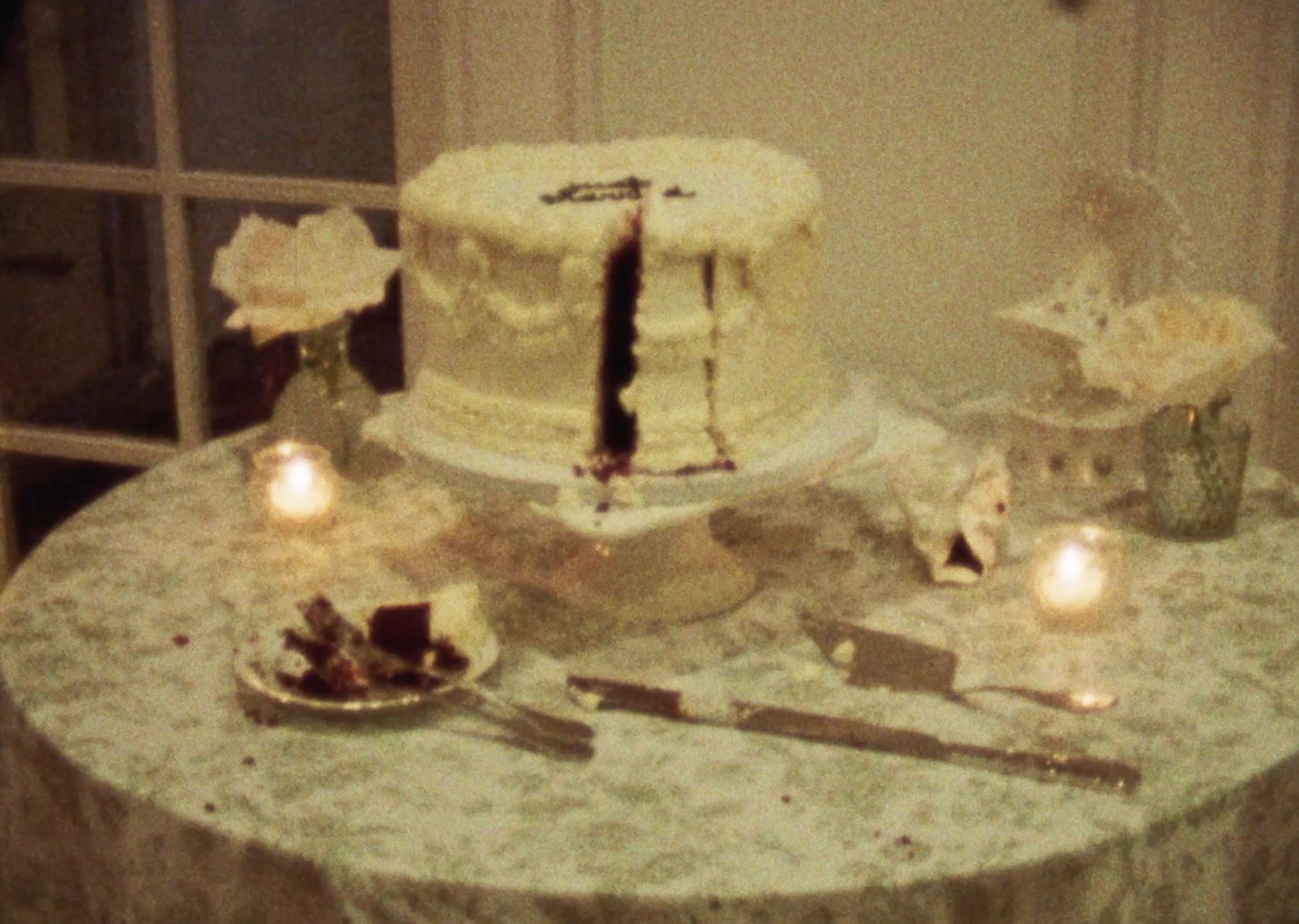 A white birthday cake with a single red candle, with a slice taken out. The cake is on a stand on a round table with a lace tablecloth. There are two lit candles, a plate with cake pieces, and some utensils on the table.