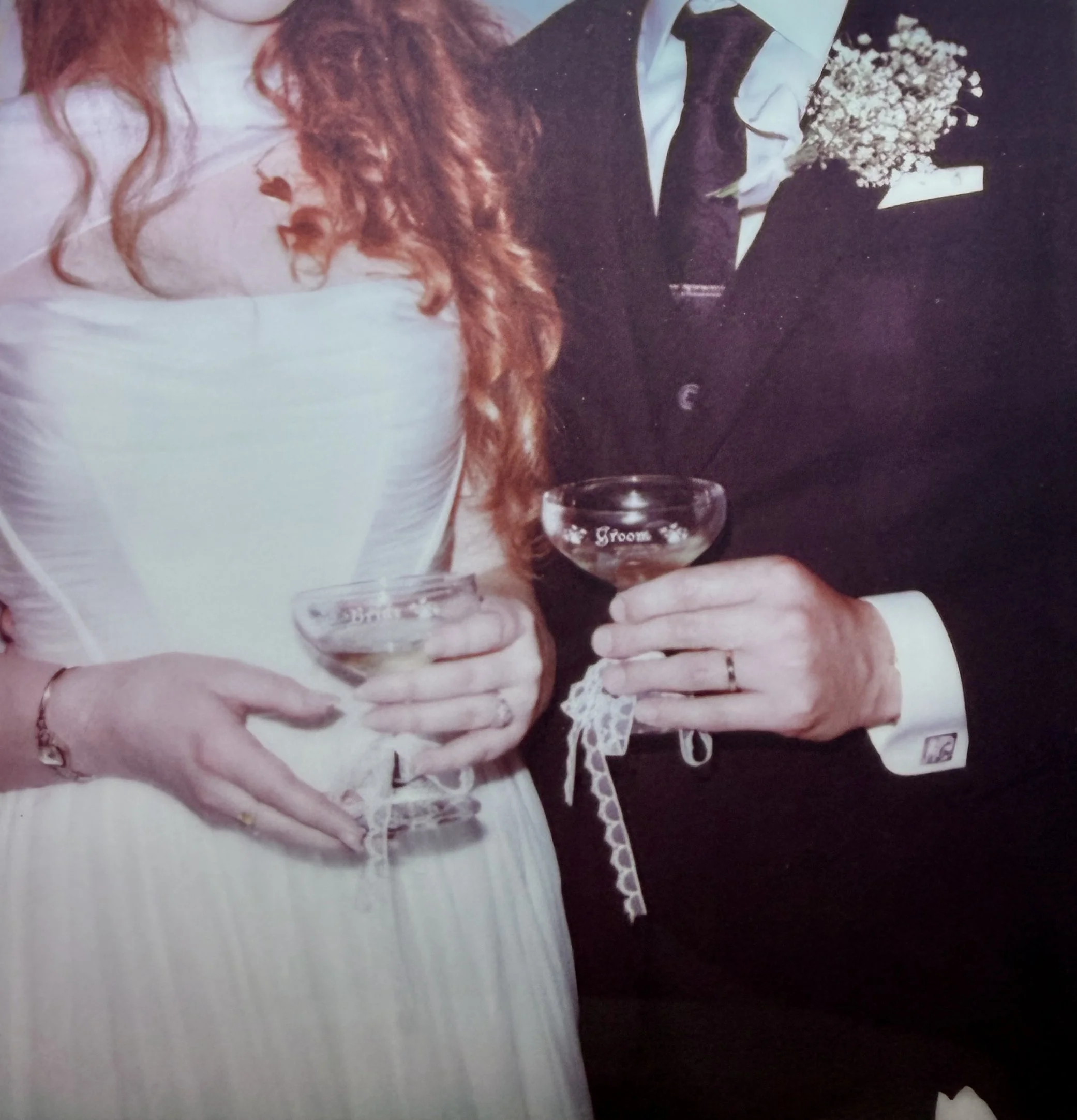 A bride and groom holding cocktails, with the bride wearing a white dress and the groom in a black suit, standing close together at their wedding celebration.