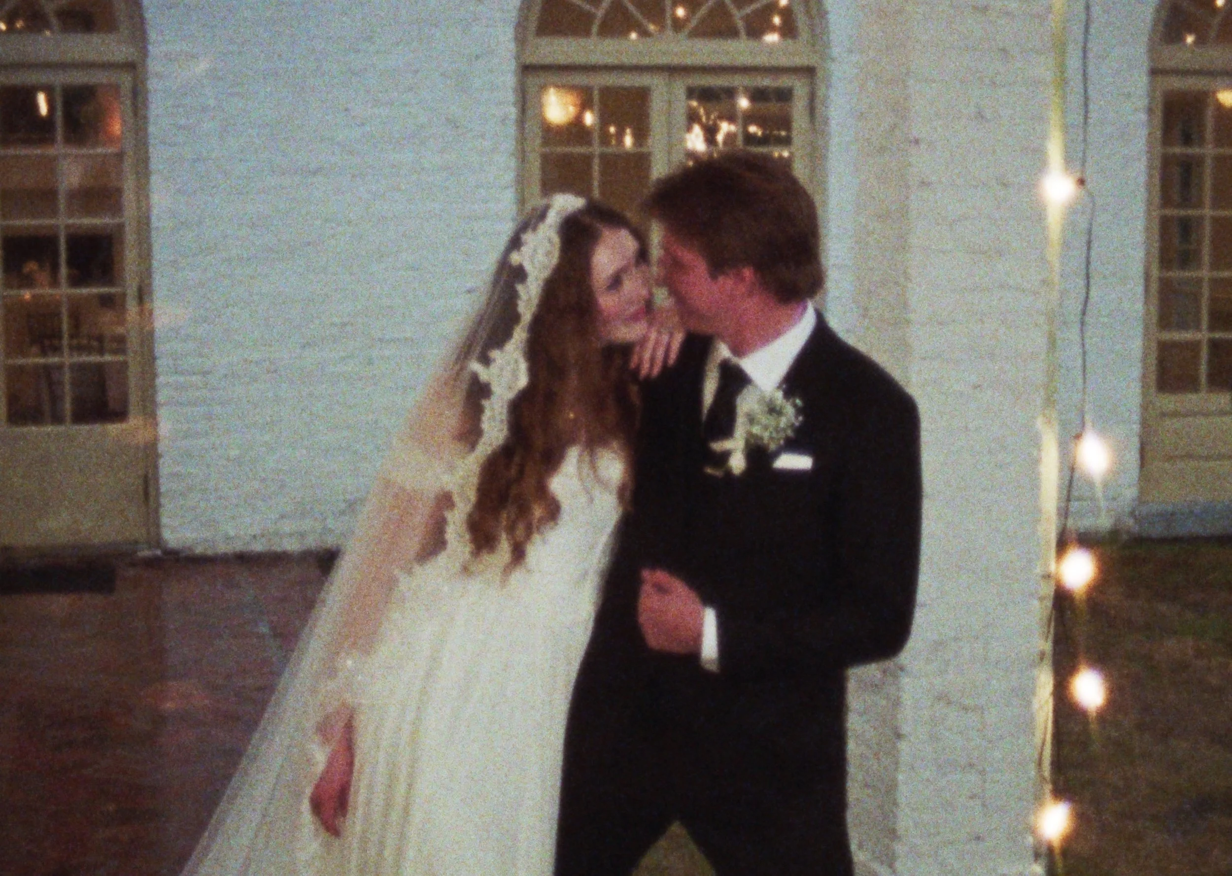 A bride and groom sharing a kiss at their wedding, with the bride wearing a white dress and veil, and the groom in a black tuxedo with a boutonniere, inside a decorated venue.