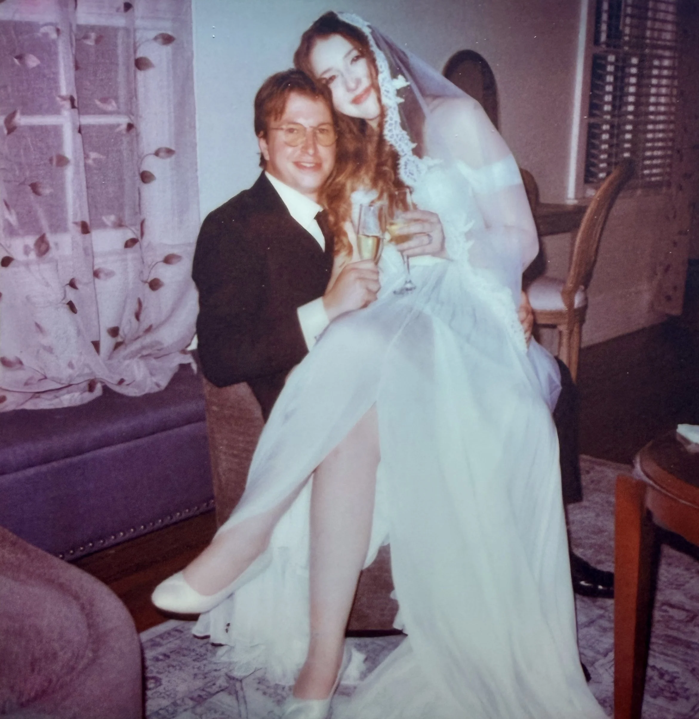 A newlywed couple celebrating with champagne in a living room, with the bride dressed in a white wedding gown and the groom in a black tuxedo.